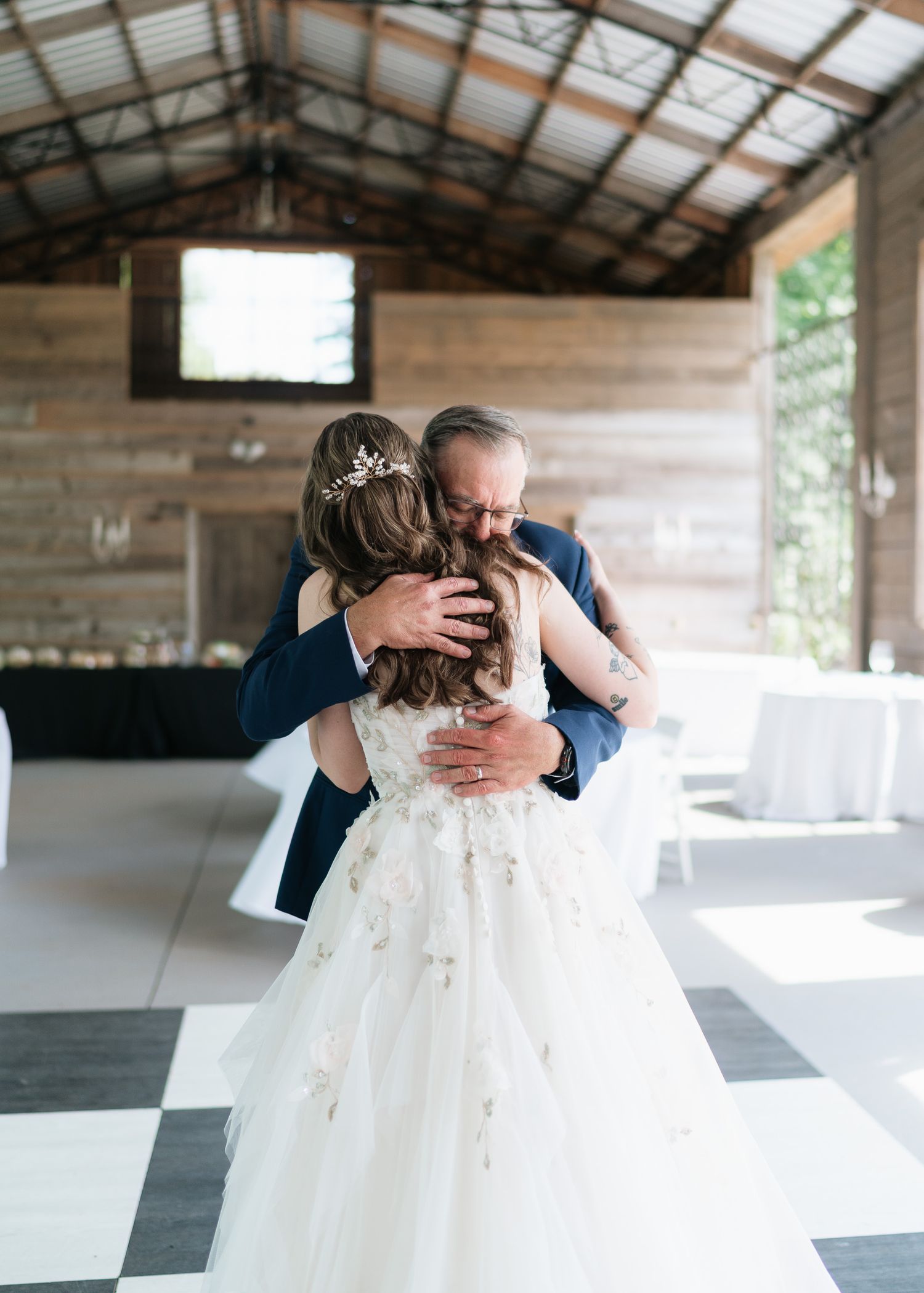Bride in floral gown shares emotional father-daughter dance hug in rustic wood-beamed reception venue.