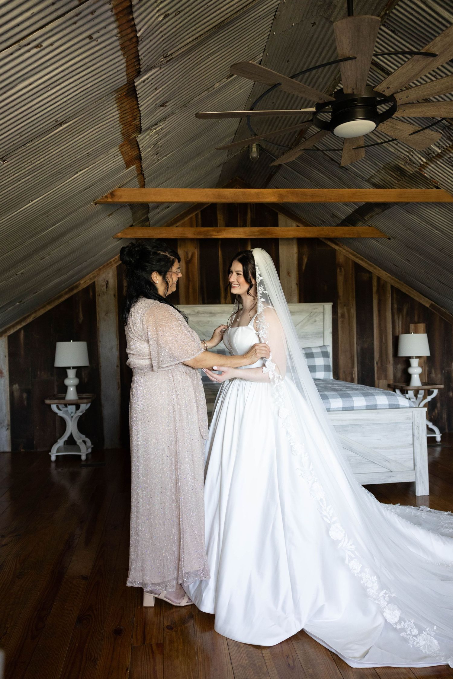 Two people in wedding attire stand together in a rustic attic space with sloped wooden ceiling and white decor.