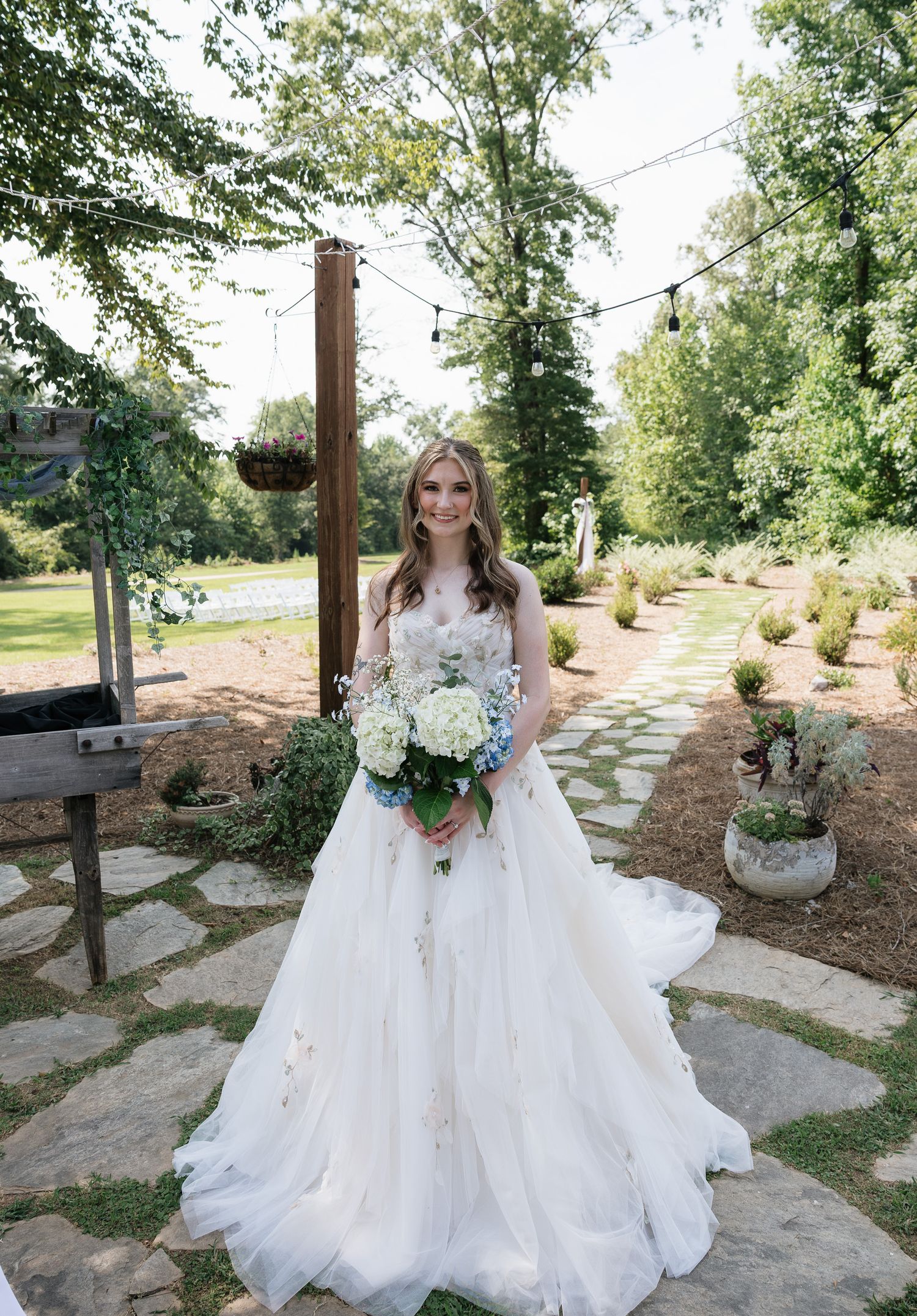 Smiling bride in floral A-line wedding gown holding white and blue hydrangea bouquet in lush garden ceremony setting.