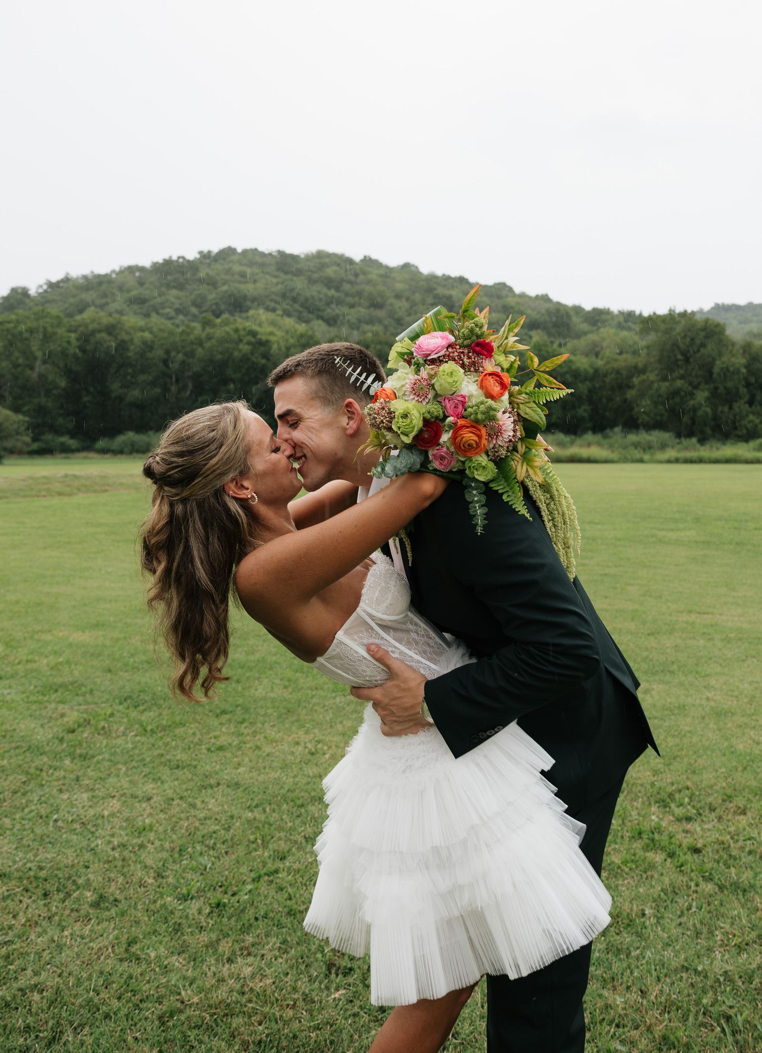 Bride and groom sharing a romantic kiss outdoors, bride holding a colorful bouquet, lush green hills in the background.