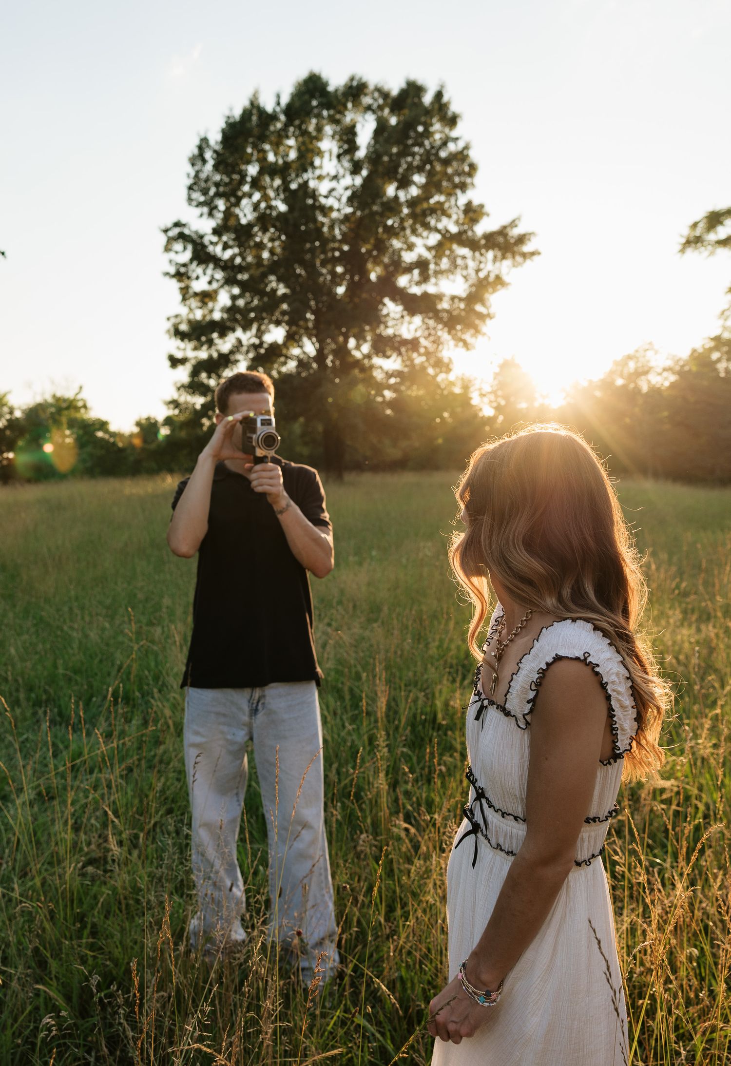 Man photographing woman in white dress at golden hour in a sunlit green field with trees in background.