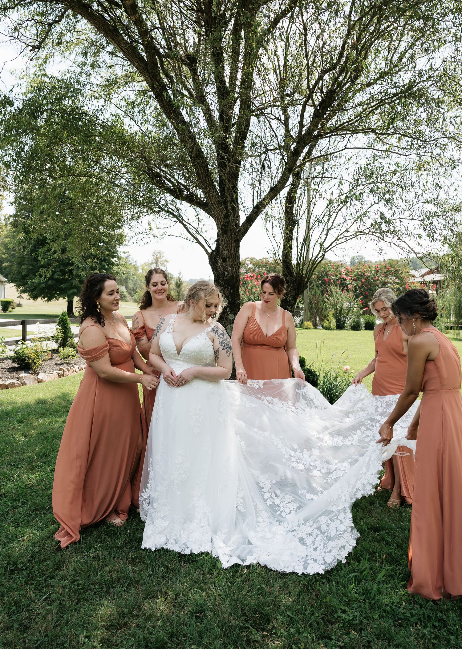 Bride in lace gown surrounded by bridesmaids in terracotta dresses arranging her train outdoors under a tree.