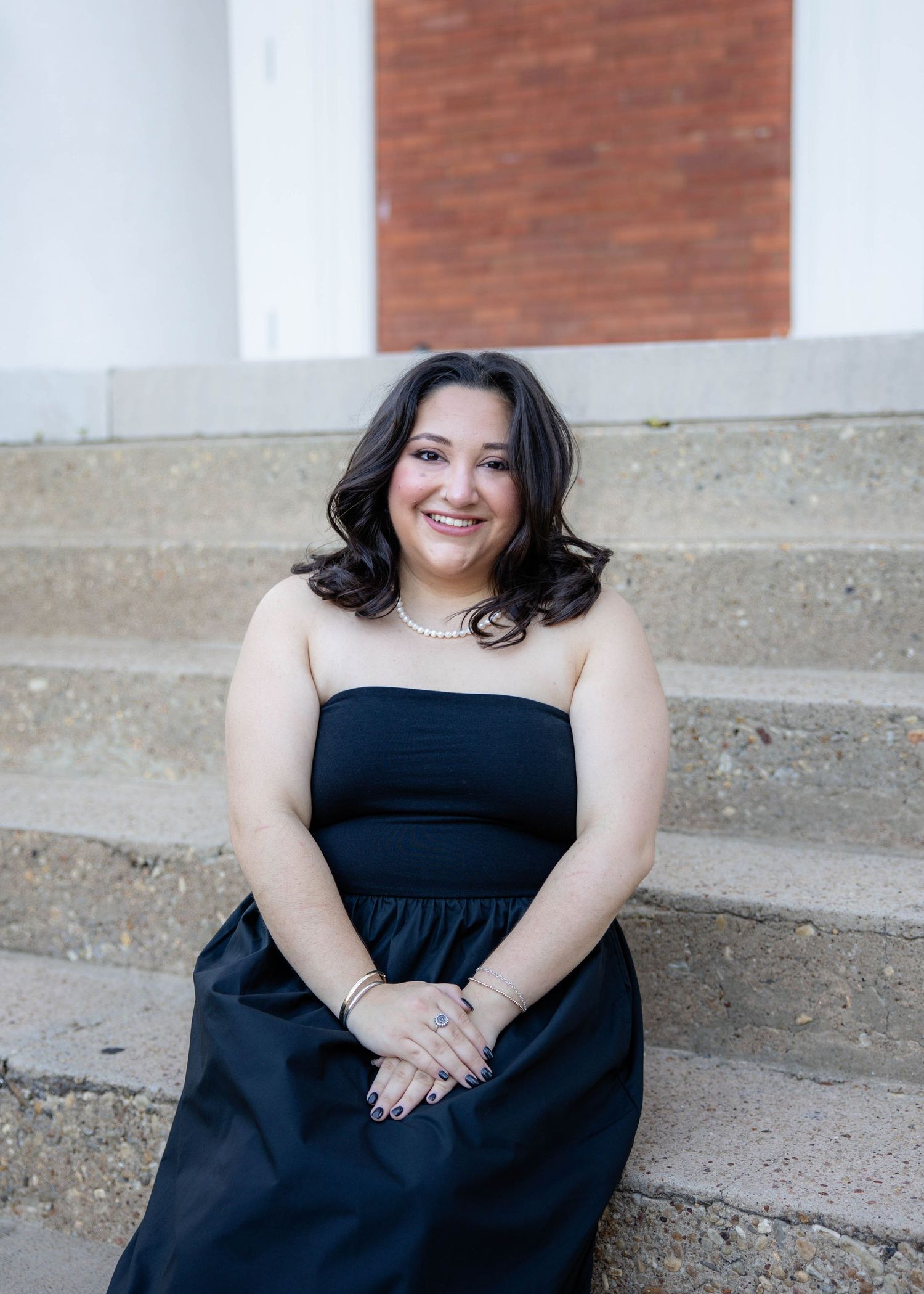 A person in a black dress sits smiling on concrete steps.