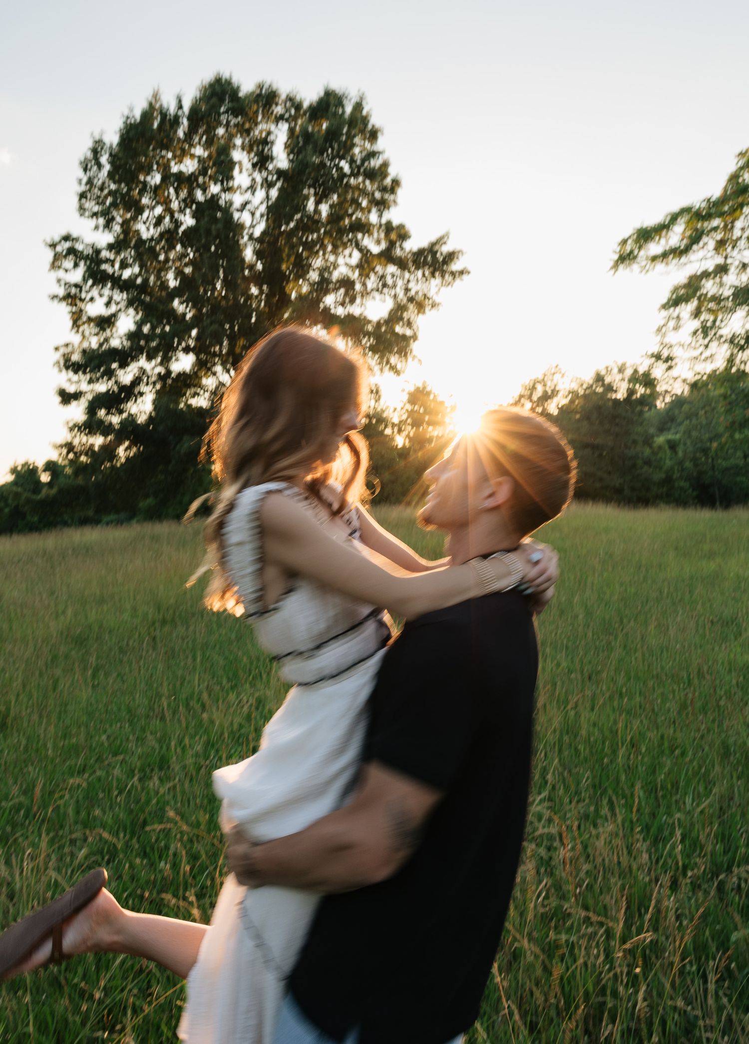 Couple sharing a romantic moment at golden hour in a lush green field, man lifting woman with sunburst behind them.