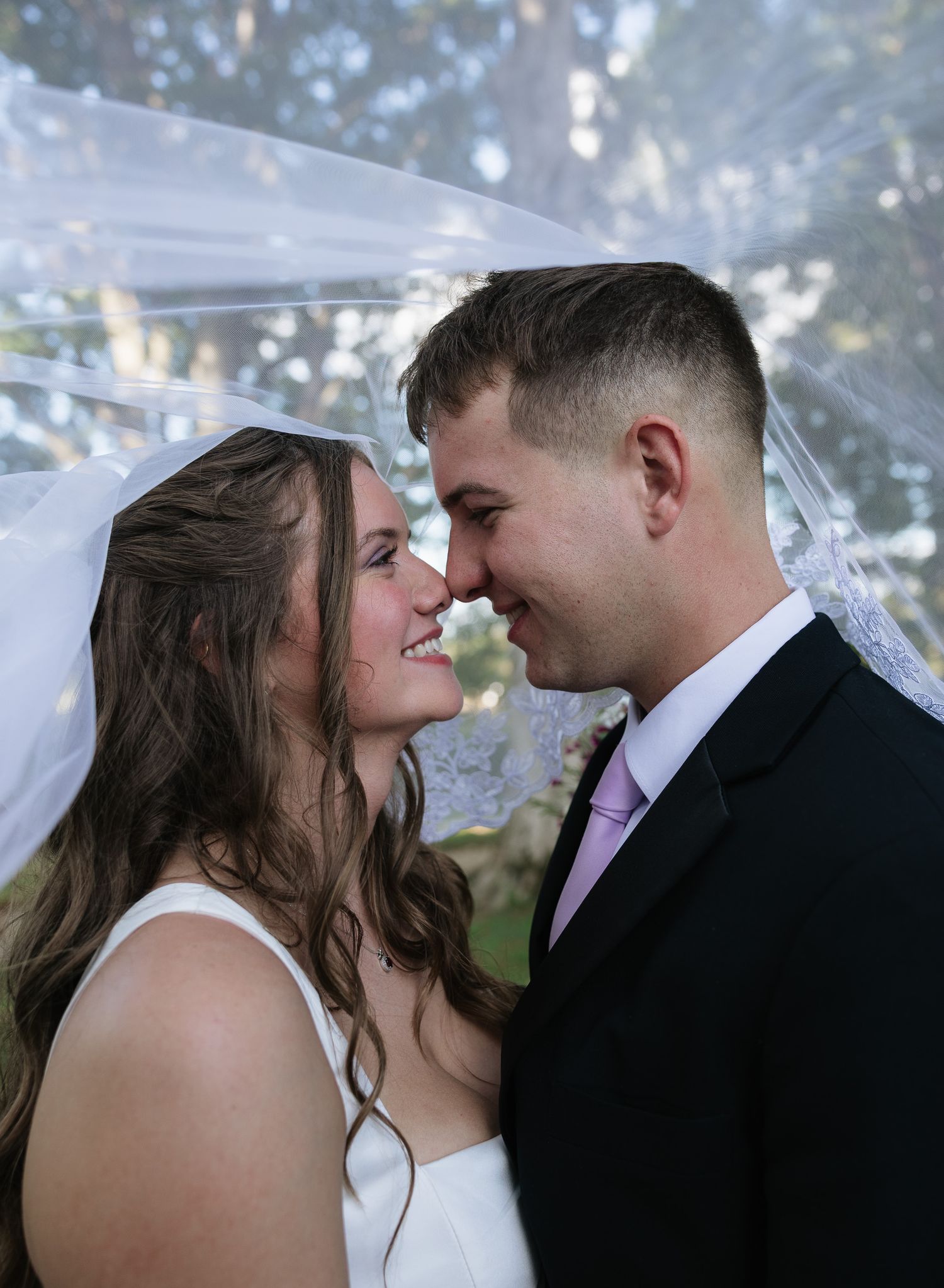 Bride and groom share a romantic moment under a veil, smiling at each other in wedding attire.