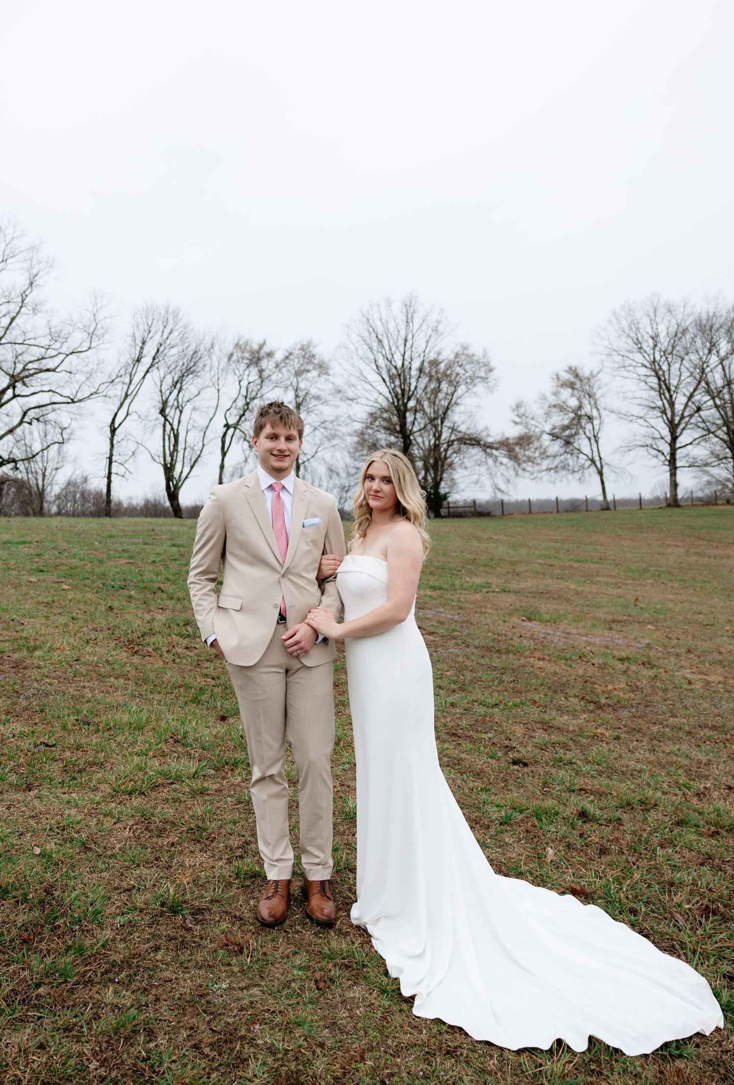 Bride in white gown and groom in tan suit pose together in a grassy field on their wedding day.