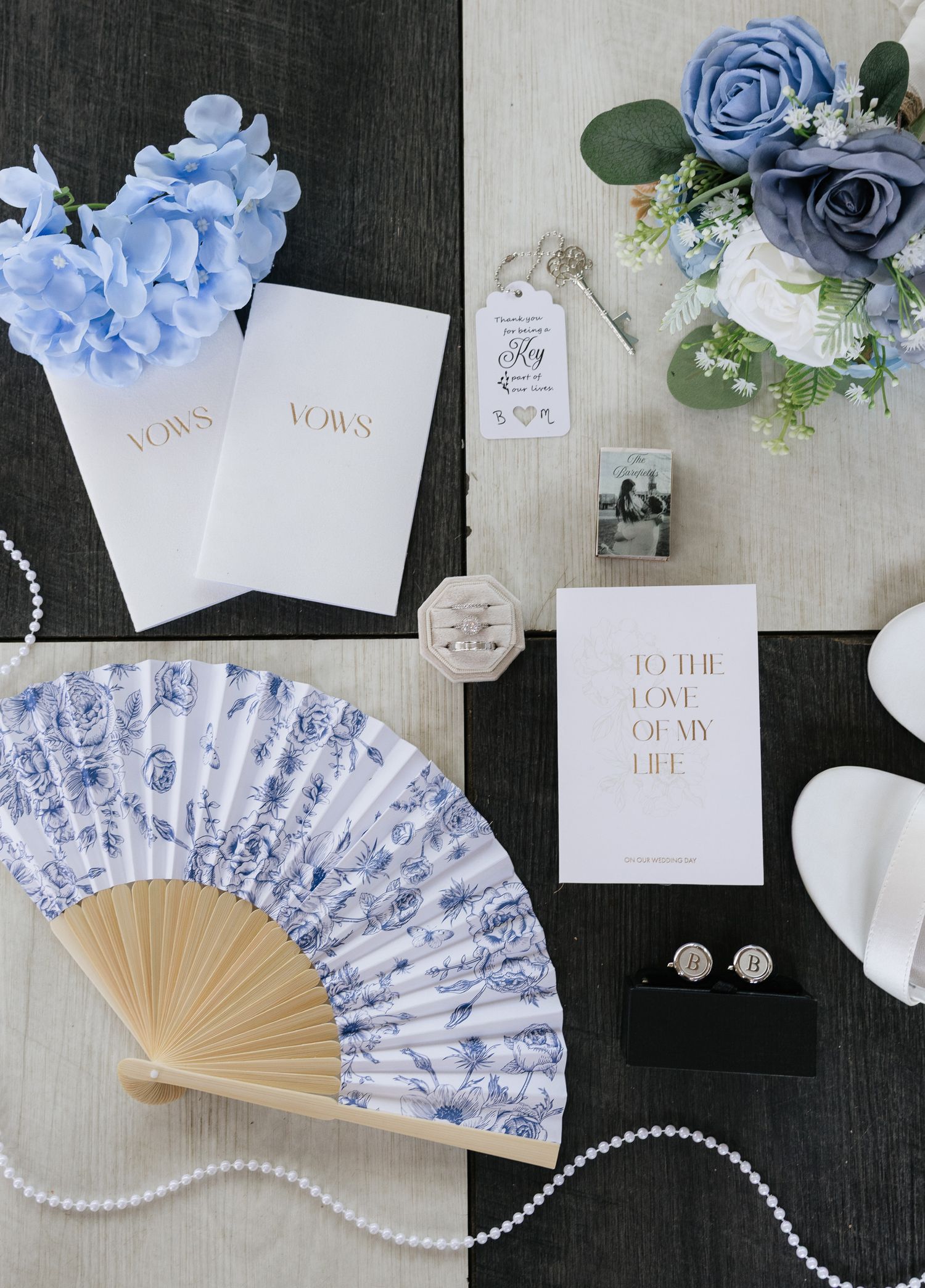 Blue and white floral fan, vow books, pearl necklace, and dusty blue roses arranged as elegant wedding flat lay.