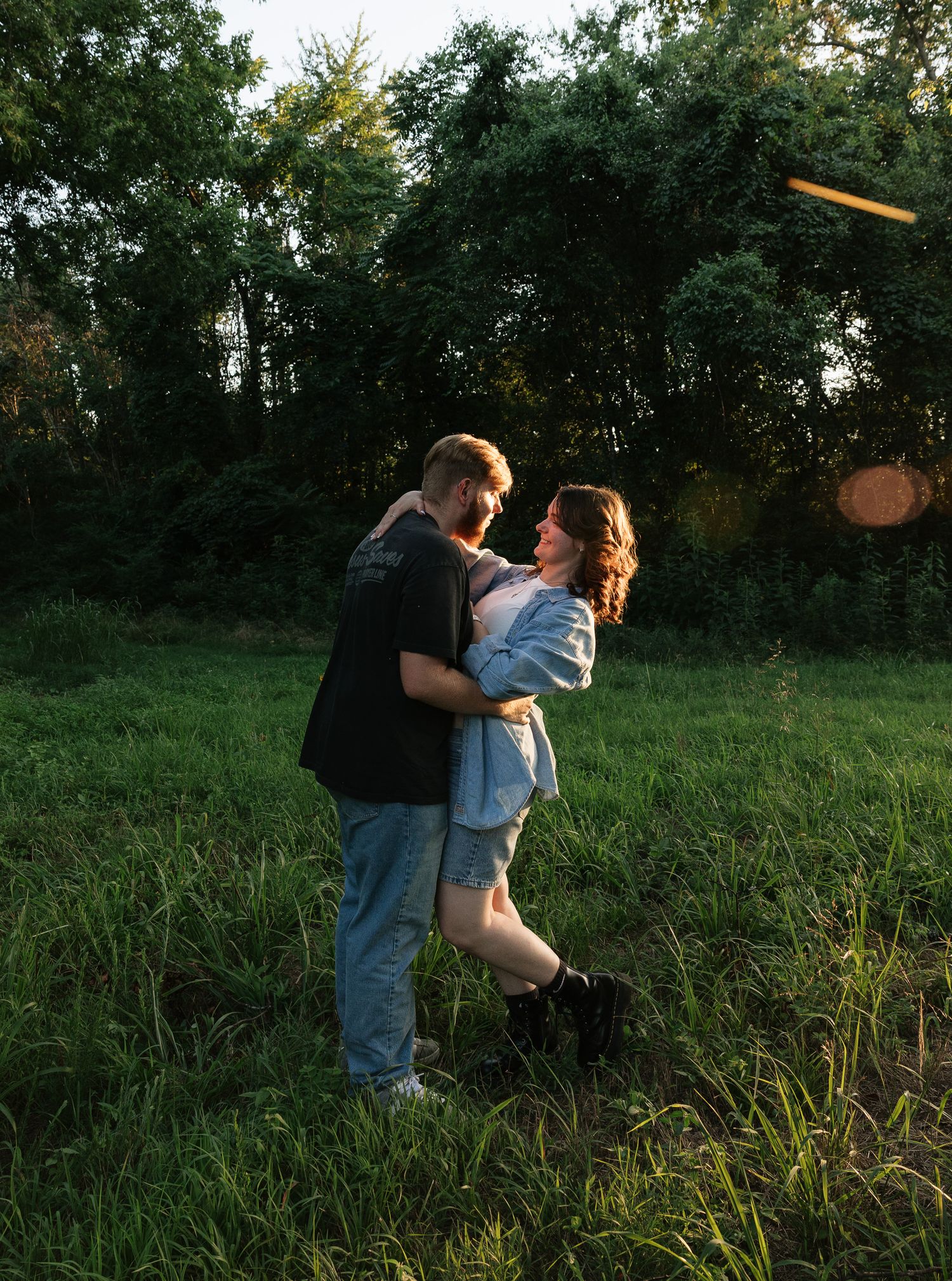 Couple embracing and dancing together in a lush green meadow surrounded by trees during golden hour sunset.