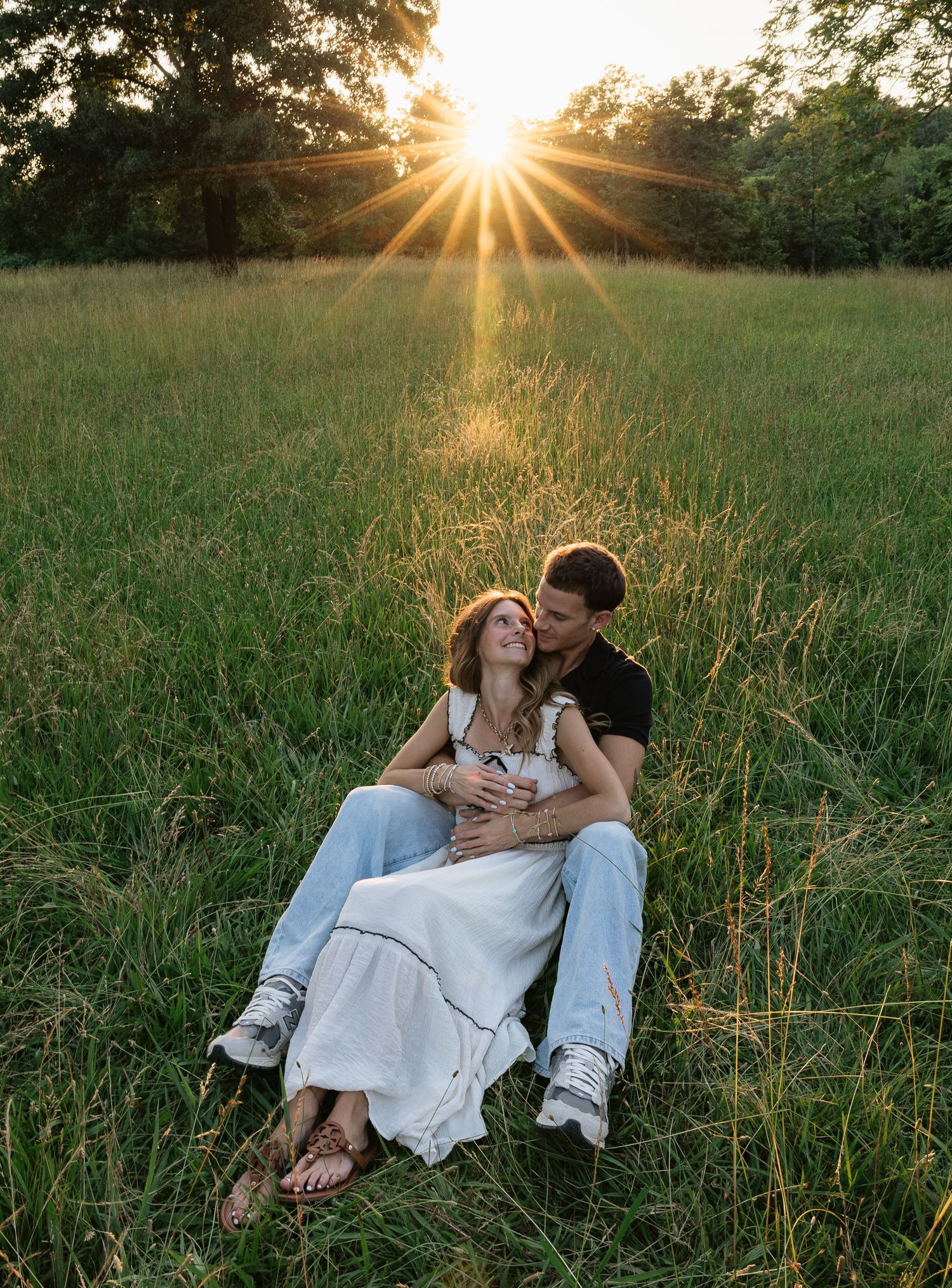 Couple sitting in a sunlit grassy field at sunset, woman in white dress leaning against man in black shirt.