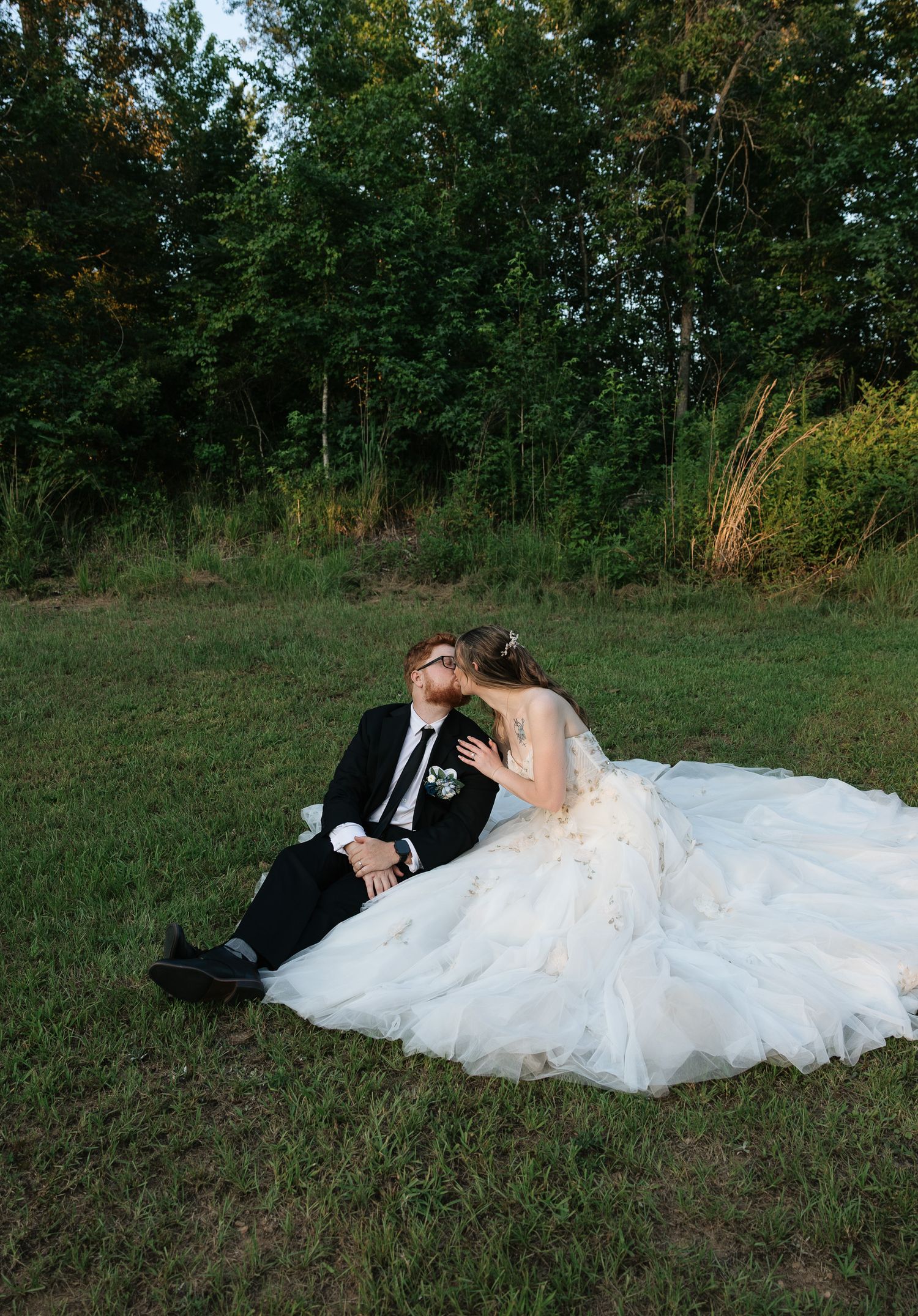 A bride and groom share a romantic kiss while sitting on a grassy lawn surrounded by lush green trees.