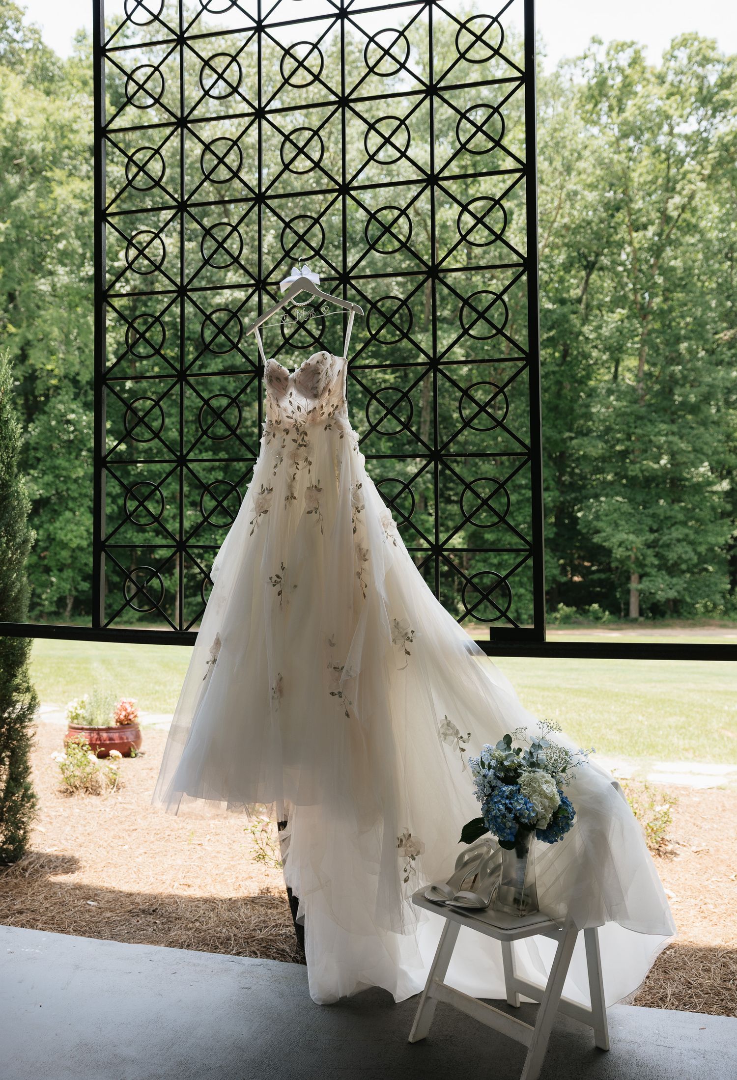 Elegant floral wedding gown hanging in front of ornate black lattice window with blue hydrangea bouquet on white stool.