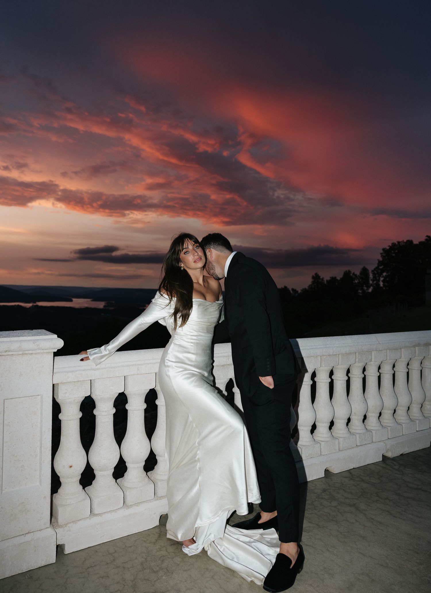 Bride in white satin gown and groom share romantic moment on balcony under dramatic red sunset sky.