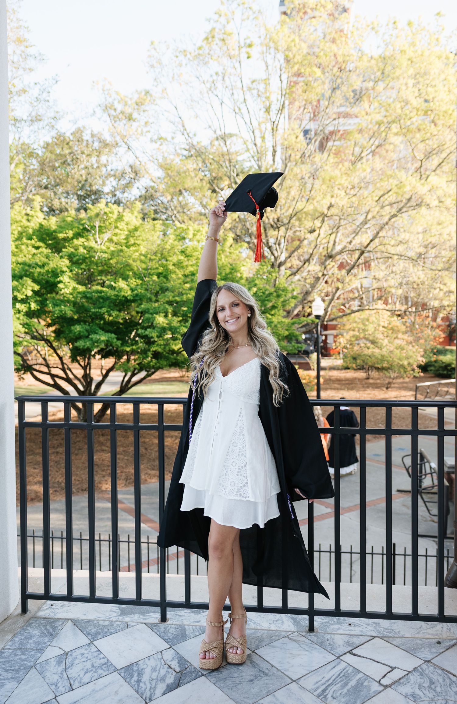 Smiling female graduate in black gown and white dress holds mortarboard aloft outdoors on a sunny day.