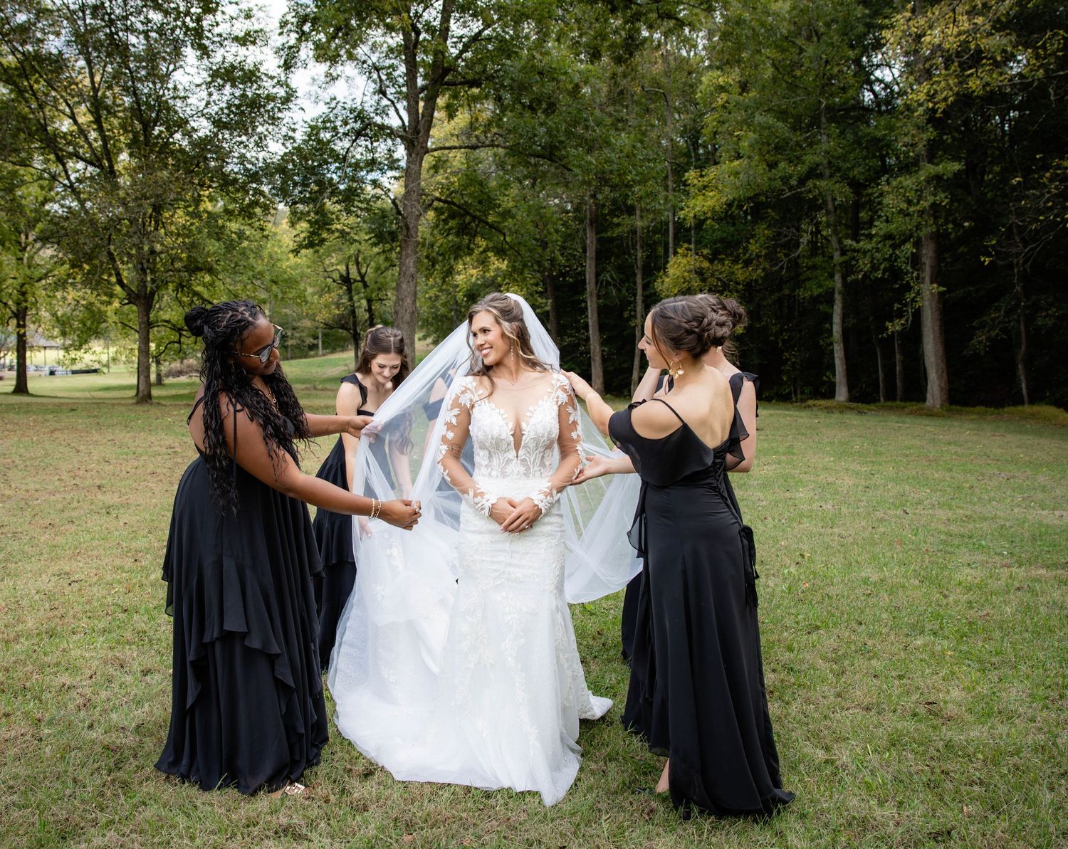 Bridesmaids in black dresses help adjust the bride's gown outdoors.