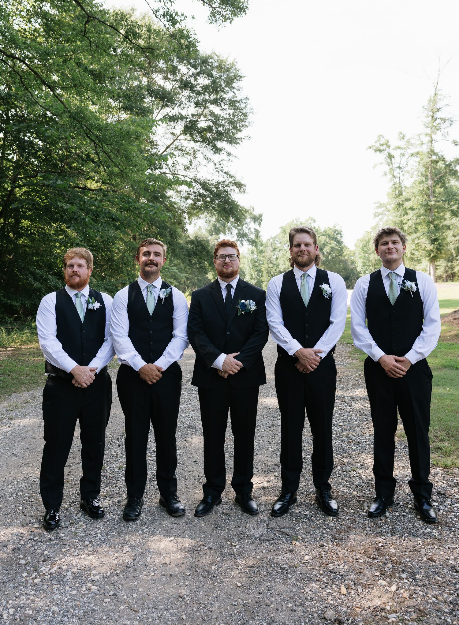 Five groomsmen in black vests and white shirts posing outdoors on a gravel path surrounded by trees.