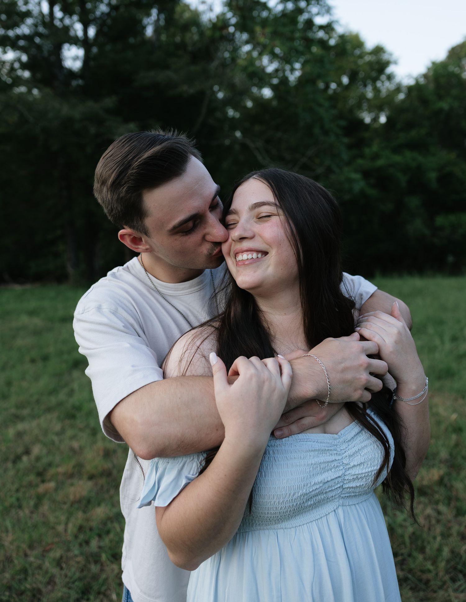 A young couple embraces outdoors, man kissing woman's cheek as she smiles, set against a lush green field.
