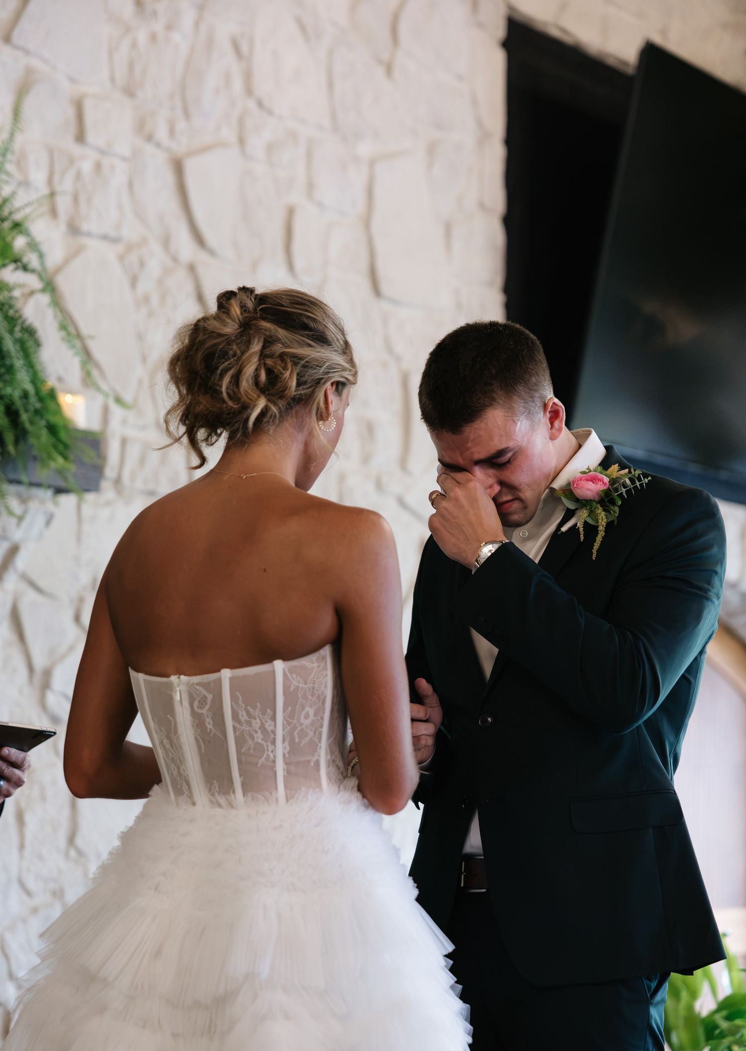 Emotional groom crying during wedding vows while bride in strapless lace corset gown stands before stone fireplace.