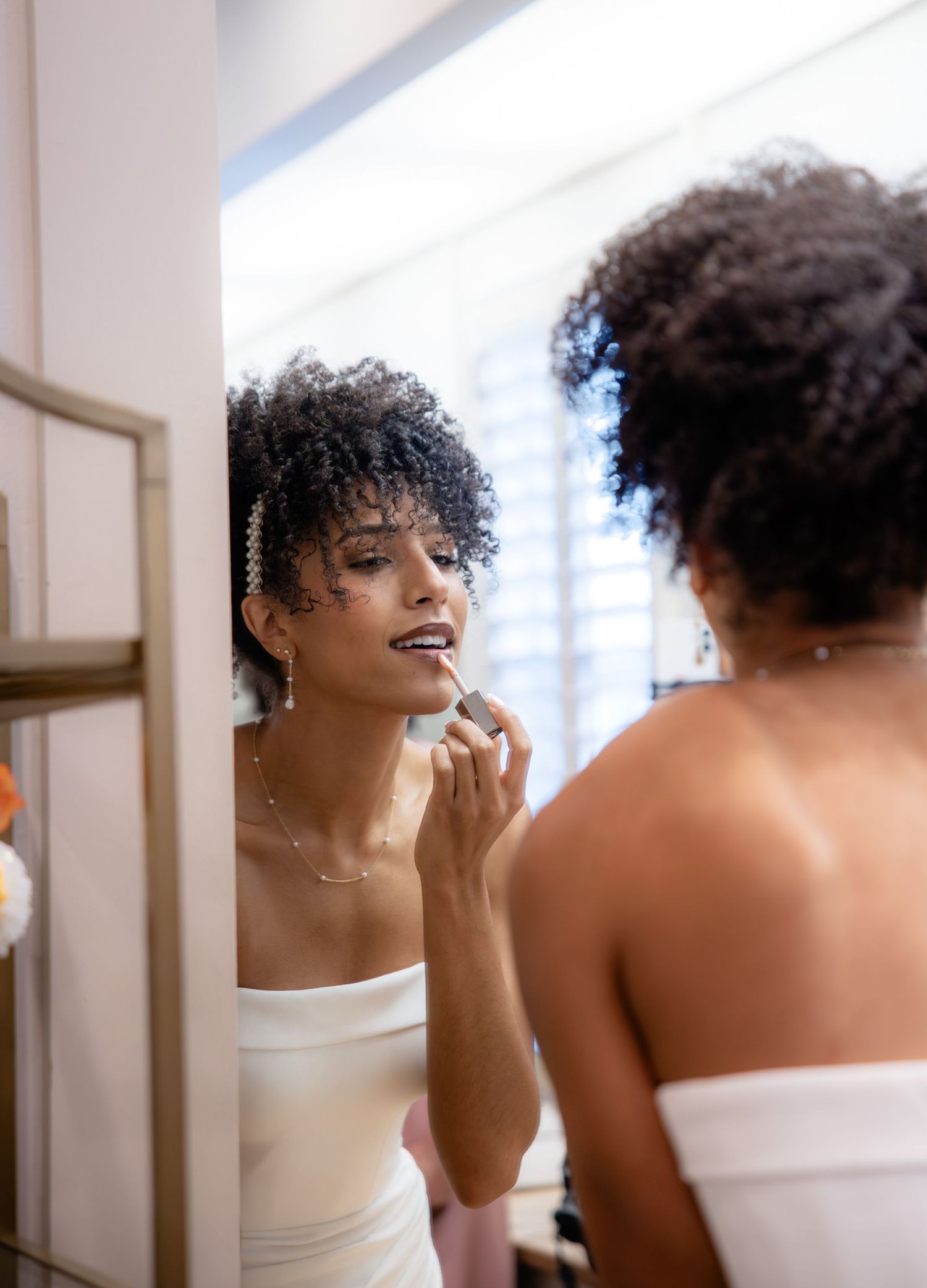 A bride in a strapless white gown applies lipstick while looking in a mirror during wedding preparations.