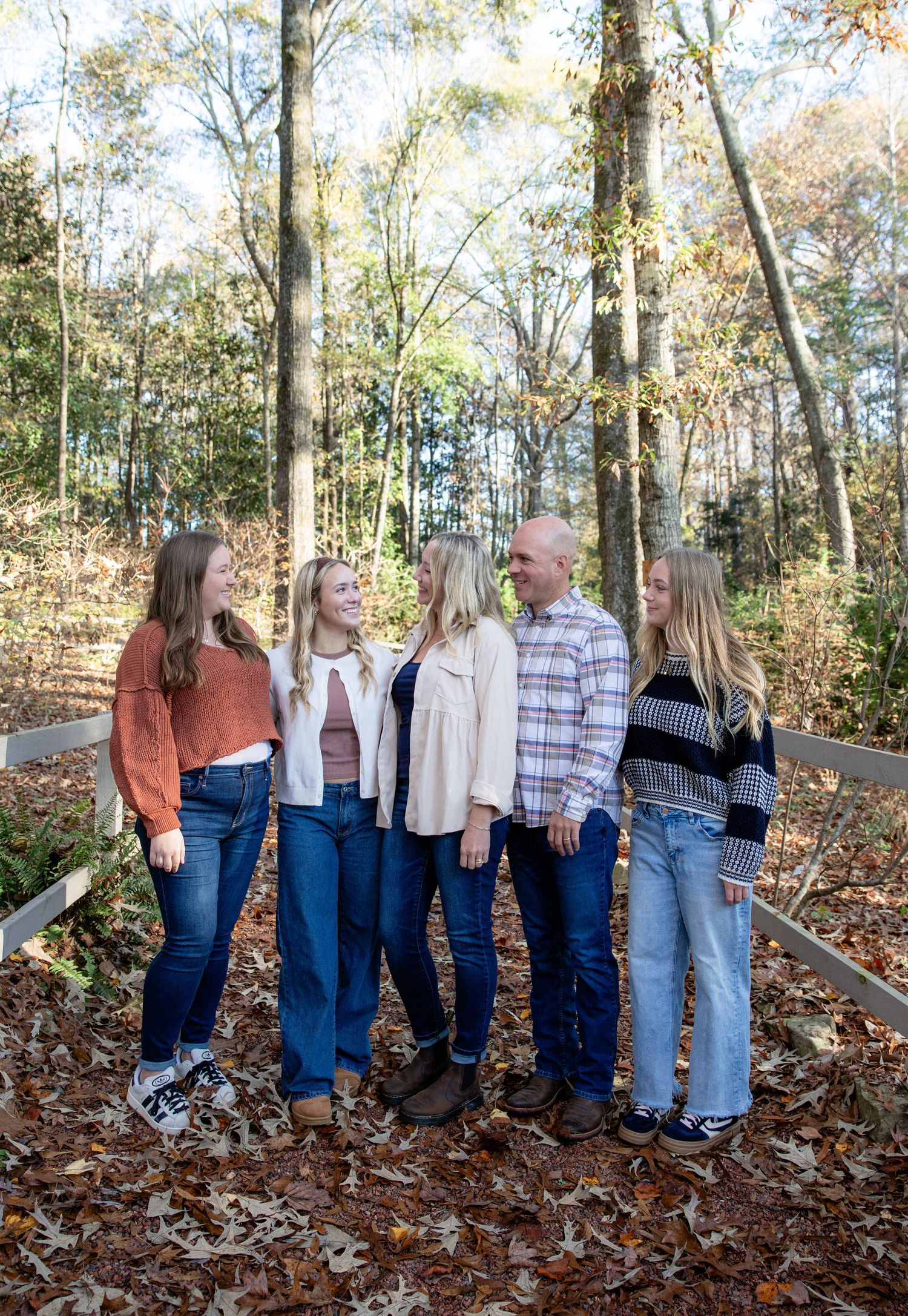 A group poses together in a wooded autumn setting wearing coordinated casual outfits in blue and neutral tones.