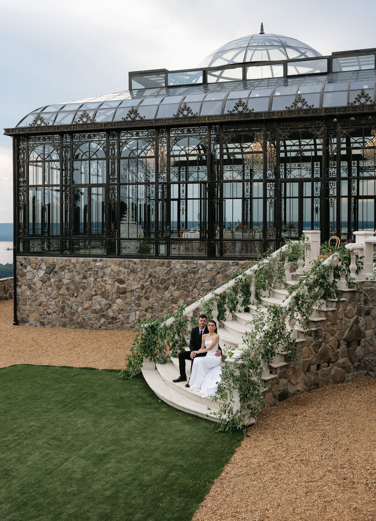 Bride and groom sit on stone steps beside a grand Victorian glass conservatory with lush greenery decor.