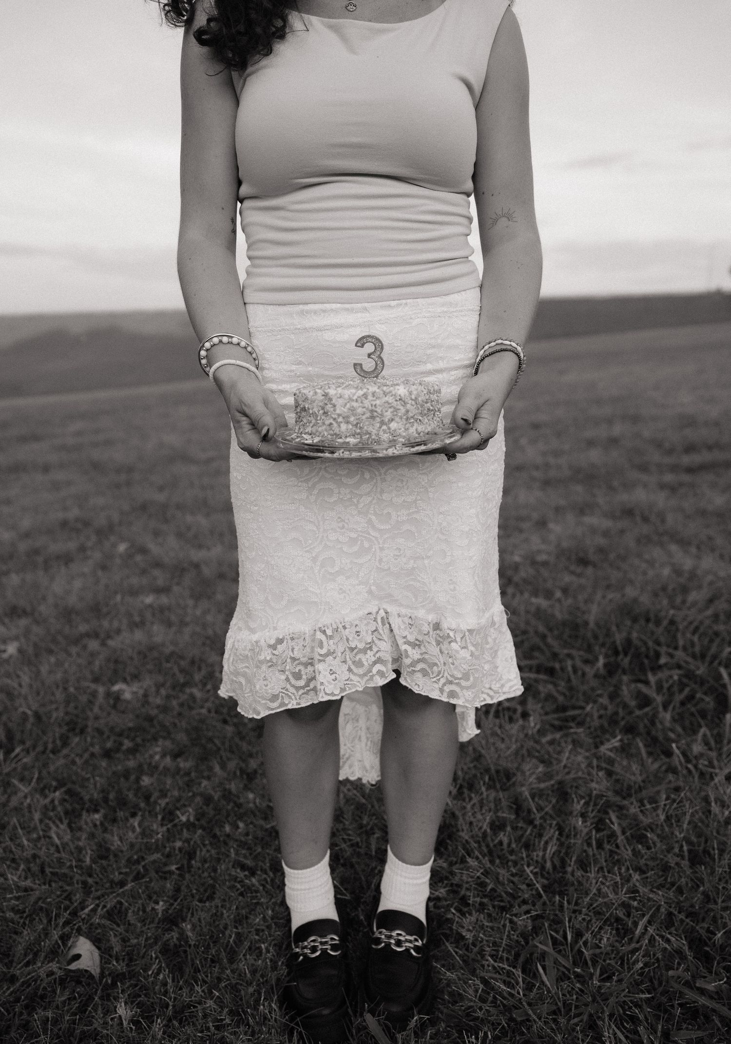 Woman in lace skirt holding a small cake with number 3 candle outdoors in a field, black and white photo.