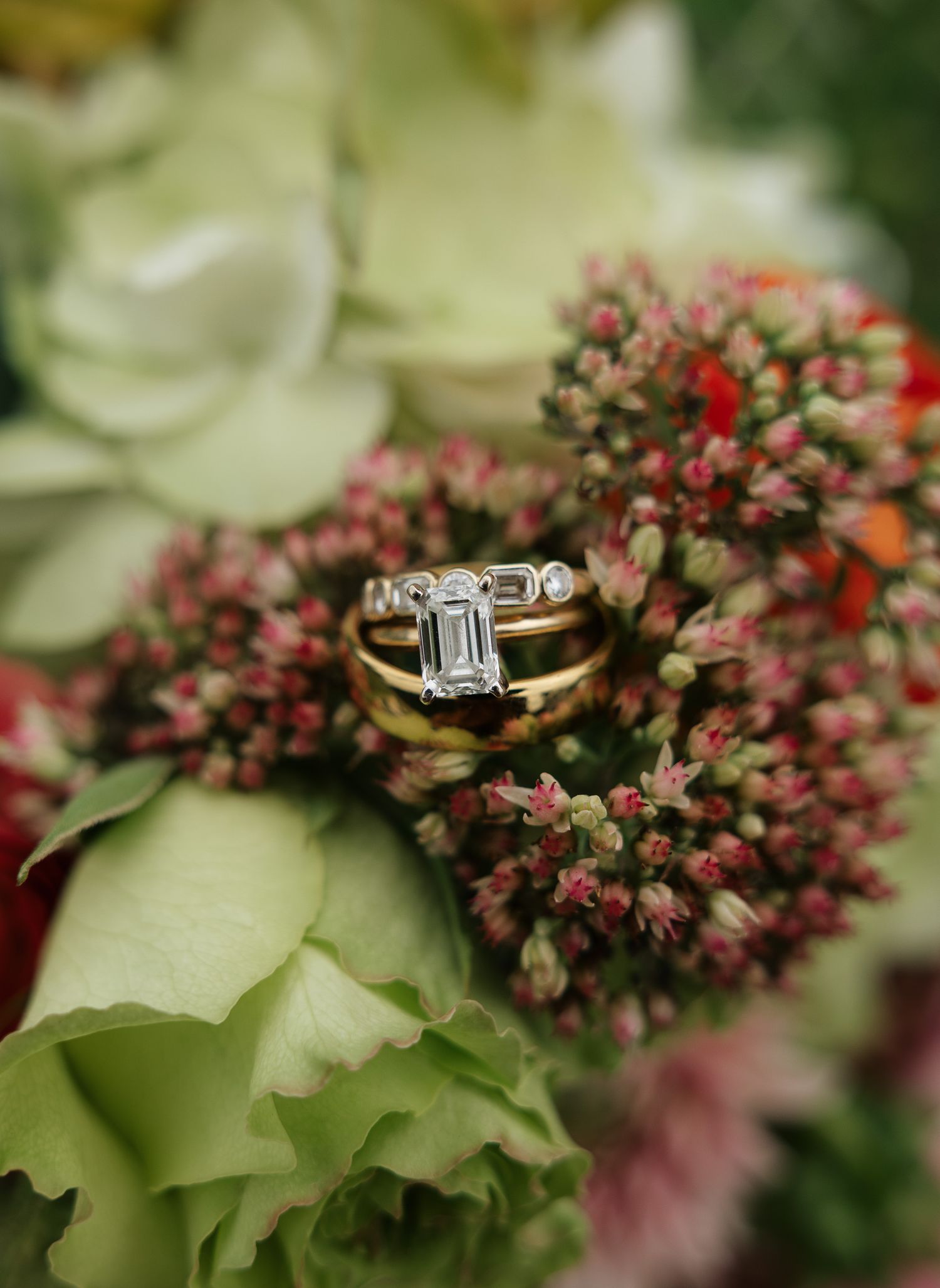 Gold engagement ring with emerald-cut diamond displayed among lush green and pink floral bouquet.