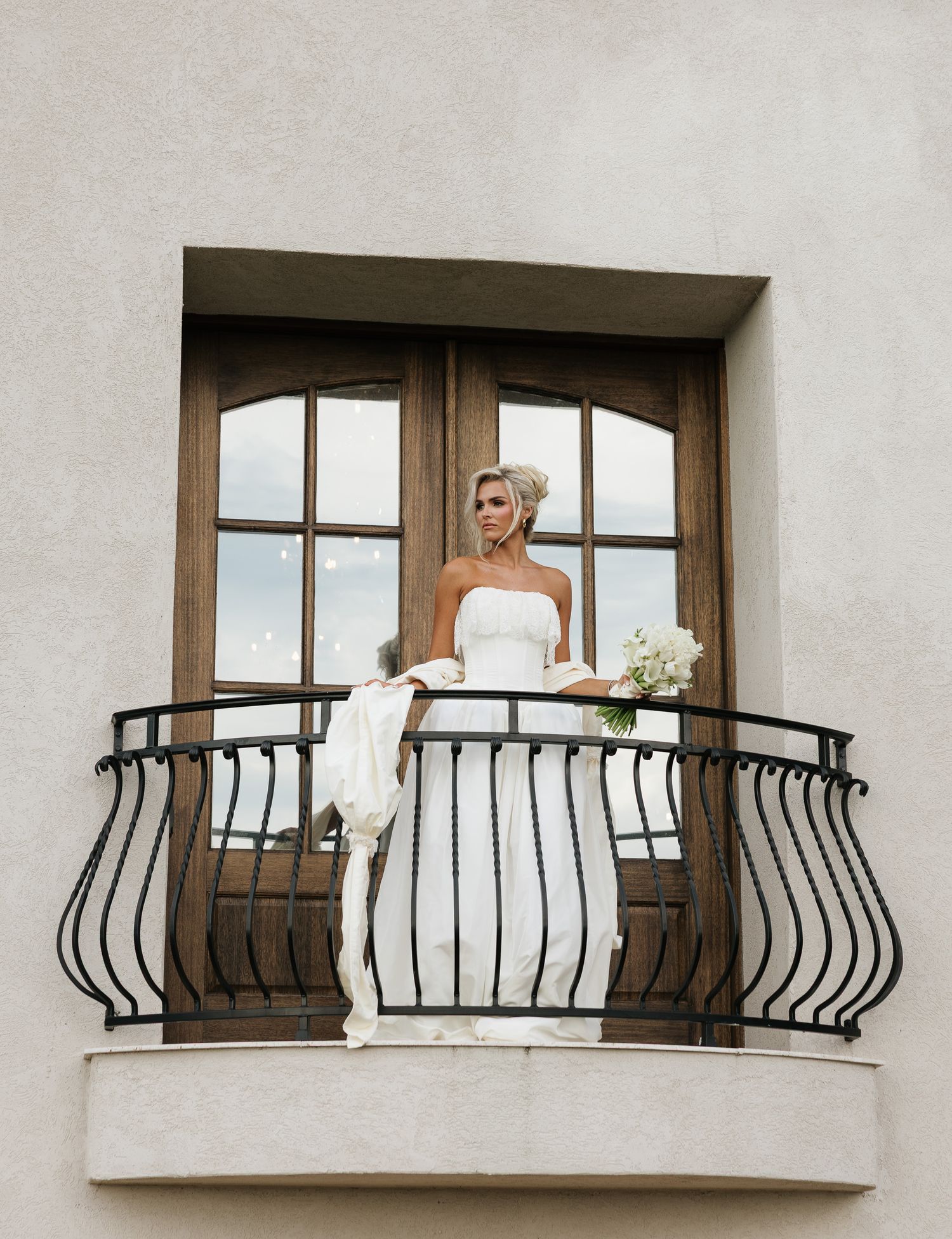 Bride in white strapless gown holding bouquet stands on ornate wrought iron balcony with wooden French doors behind her.