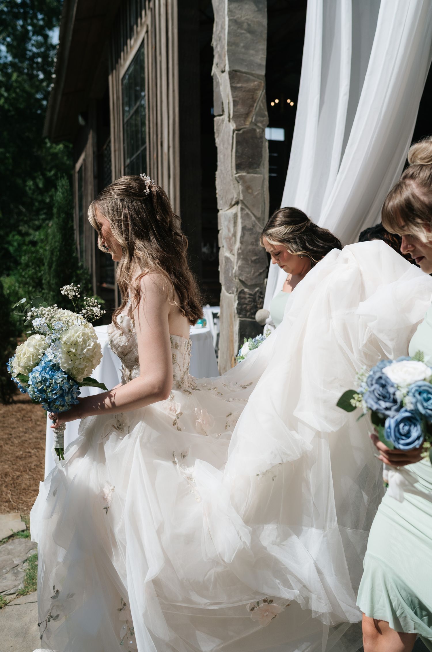 Bride in white tulle gown holding blue hydrangea bouquet walks with bridesmaid carrying her train outdoors.
