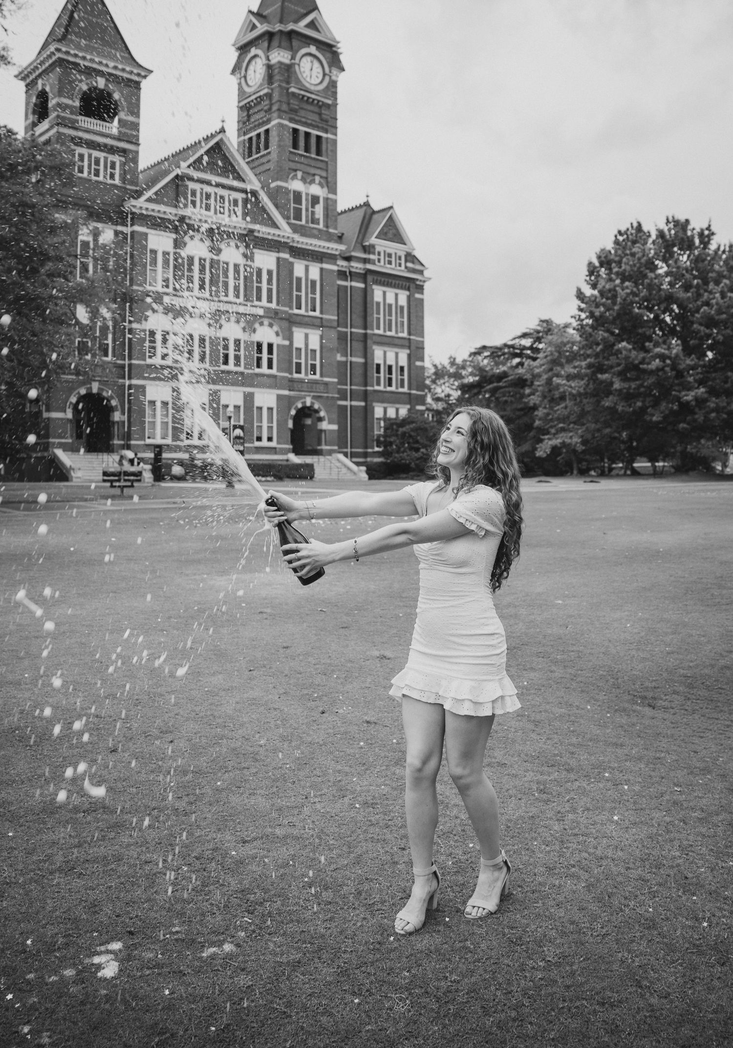 Black and white photo of a woman in a white dress popping champagne outside a brick university building.