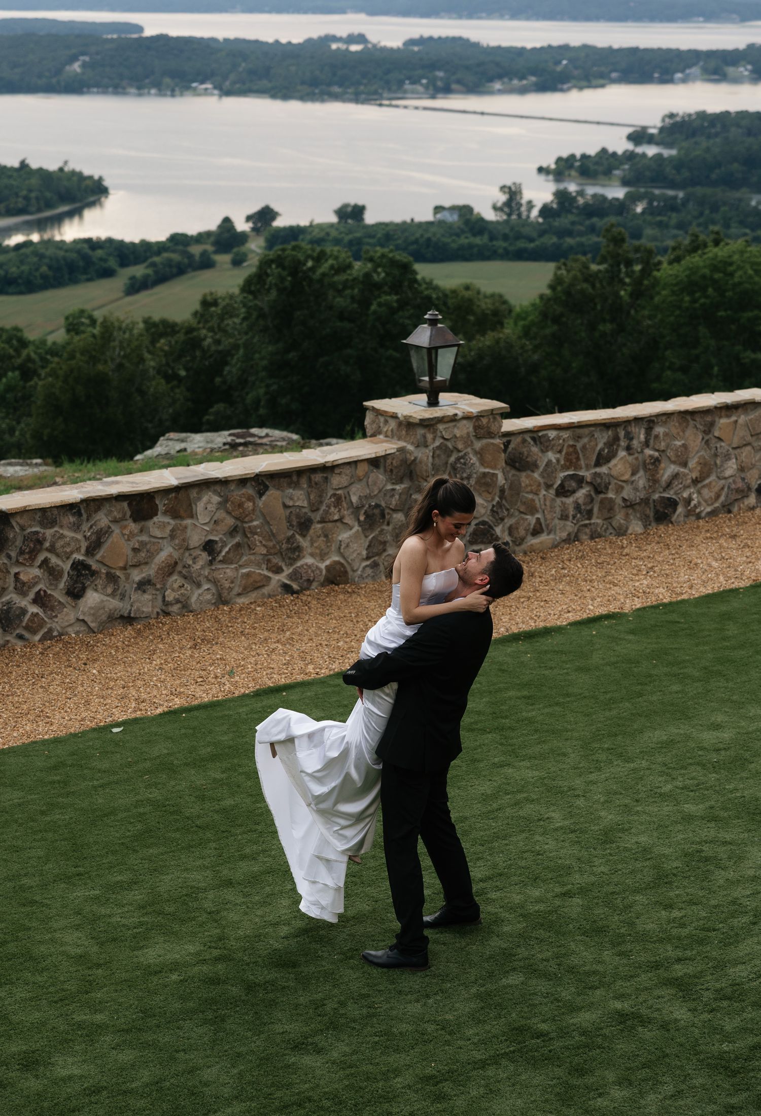 Groom lifting bride in a romantic kiss on a hilltop lawn with scenic river views behind them.