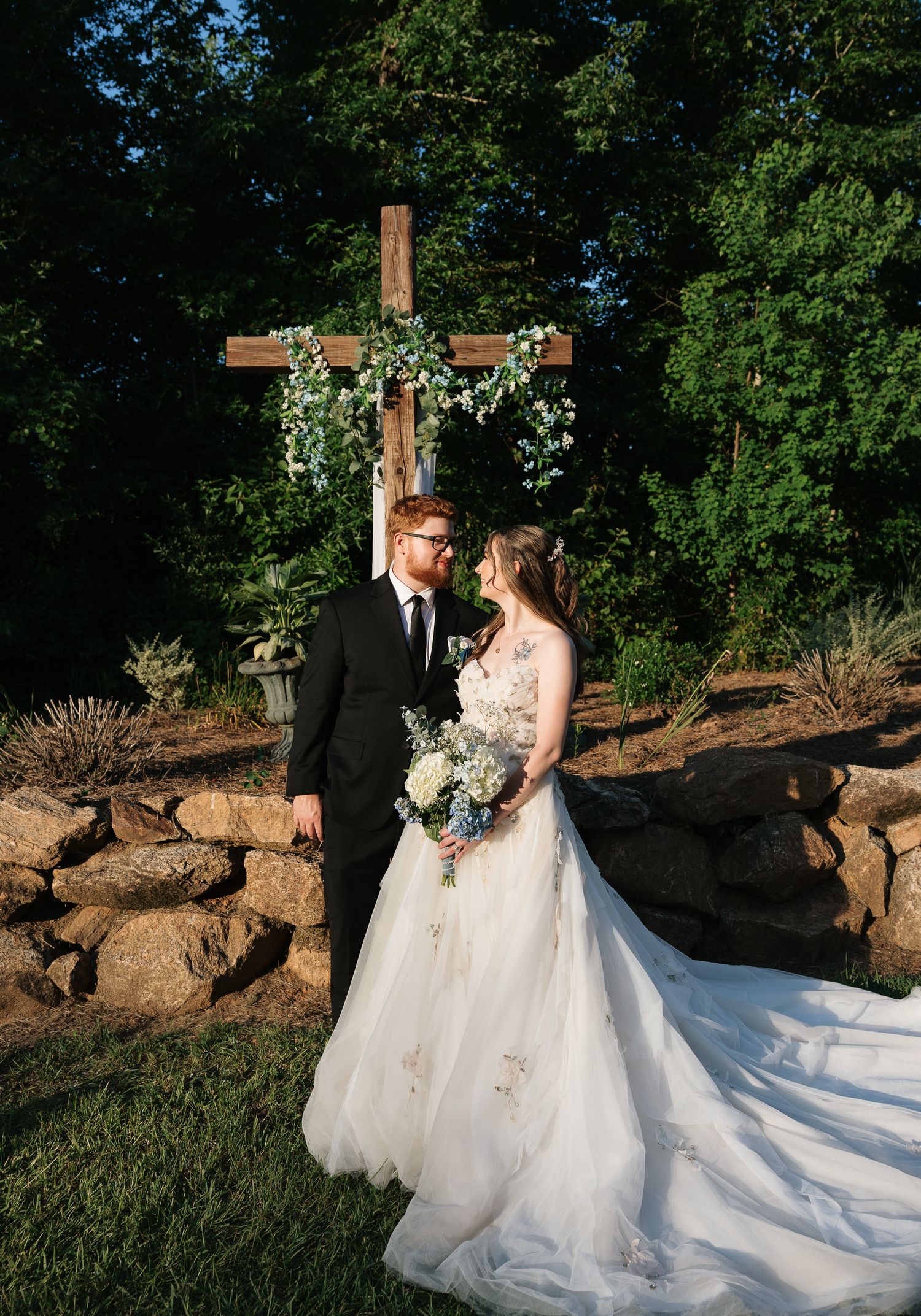 Bride and groom stand before a wooden cross adorned with greenery at an outdoor wedding ceremony.