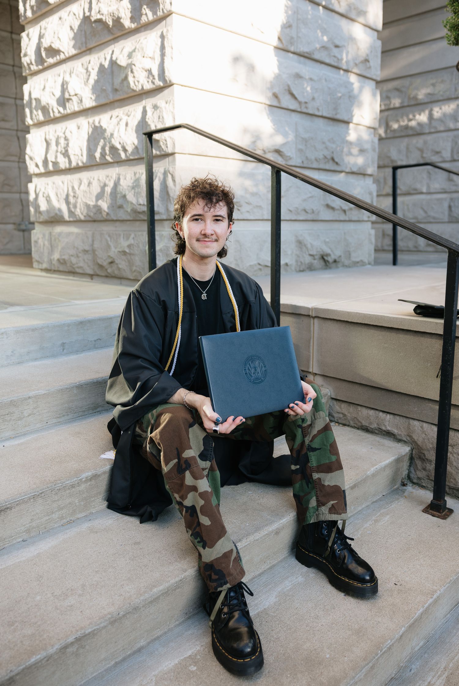 Young graduate in black gown and camo pants holds diploma on stone steps, smiling proudly at camera.