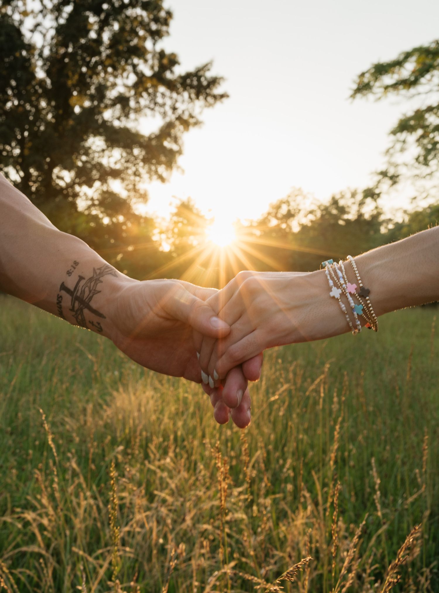Two people holding hands in a sunlit field at sunset, one with a tattoo and the other wearing colorful bracelets.