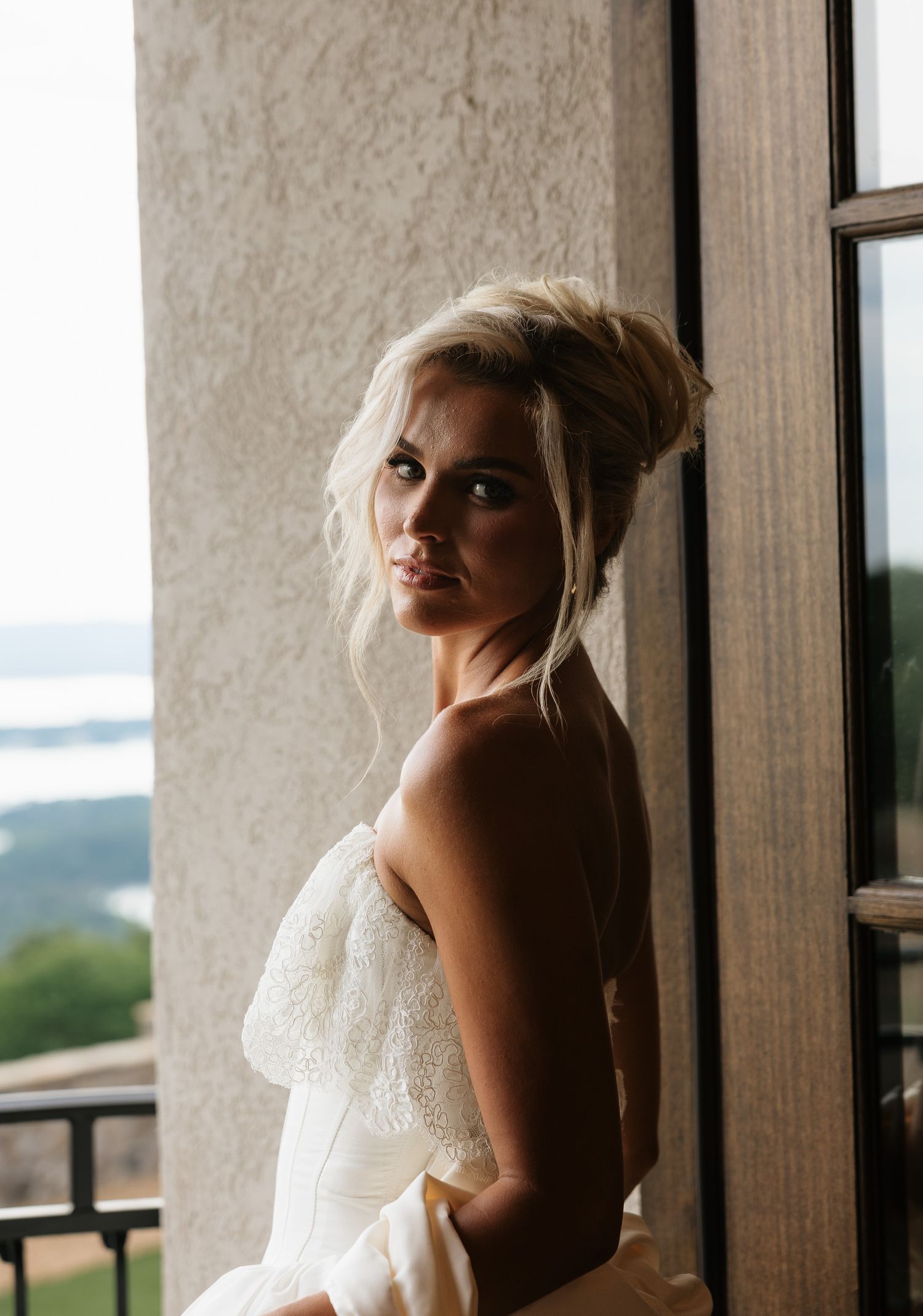 Elegant bride in strapless lace wedding gown poses by a window with scenic outdoor views, blonde updo hairstyle.