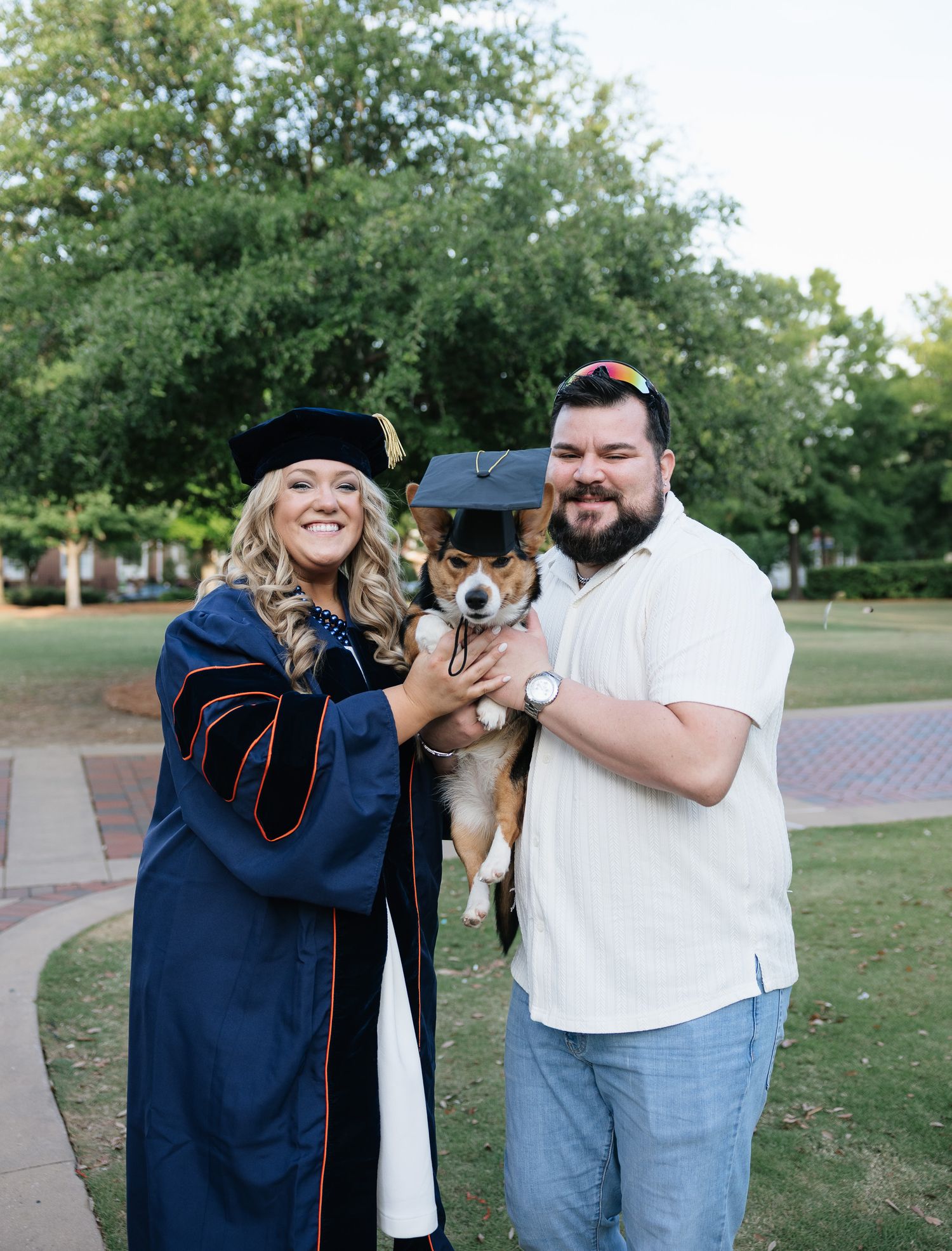 A woman in doctoral graduation regalia poses with a man and their corgi dog wearing a tiny graduation cap outdoors.