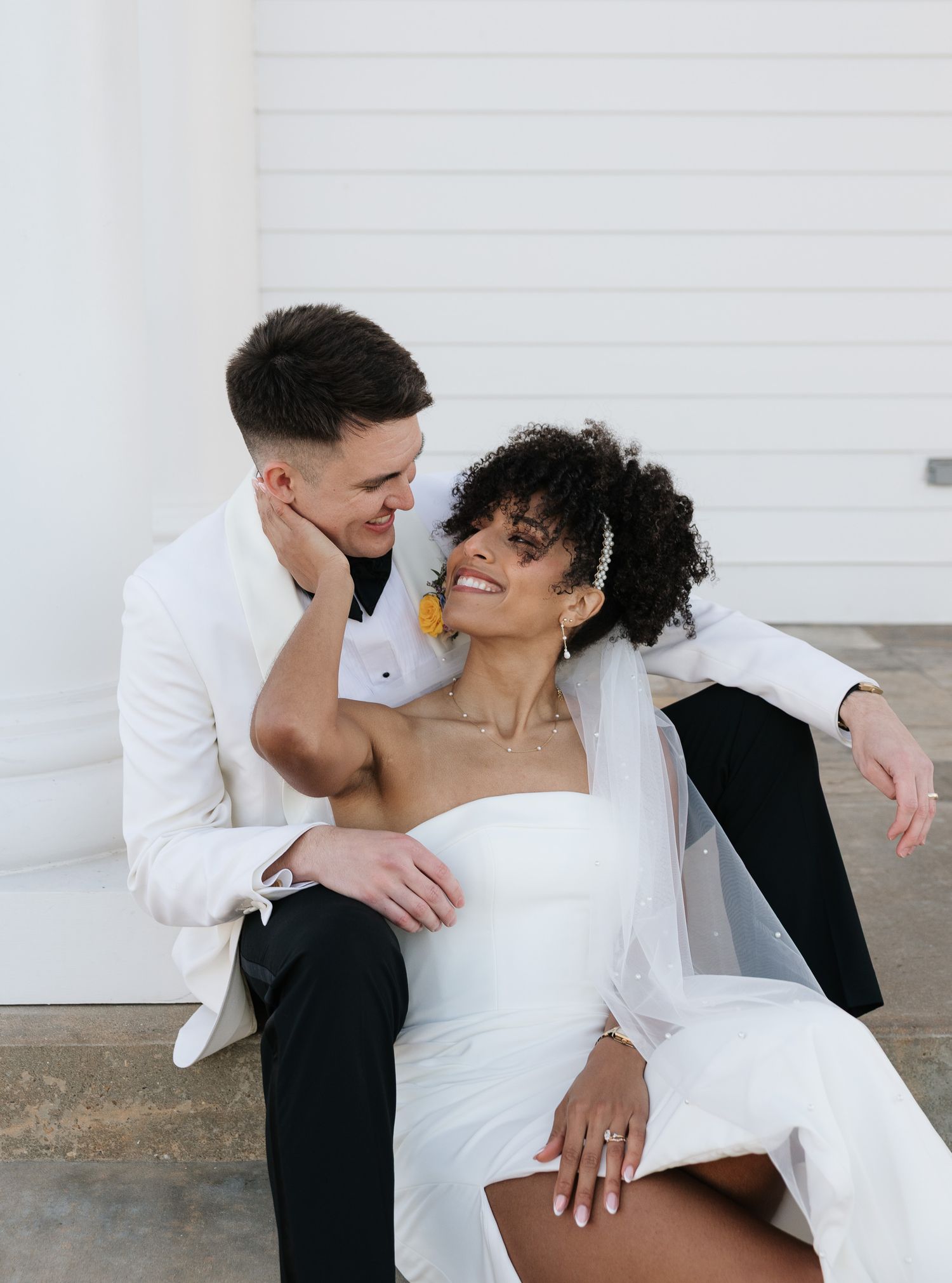 A couple in wedding attire sits together, the groom in a white suit and bride in a strapless white dress, sharing a loving gaze.