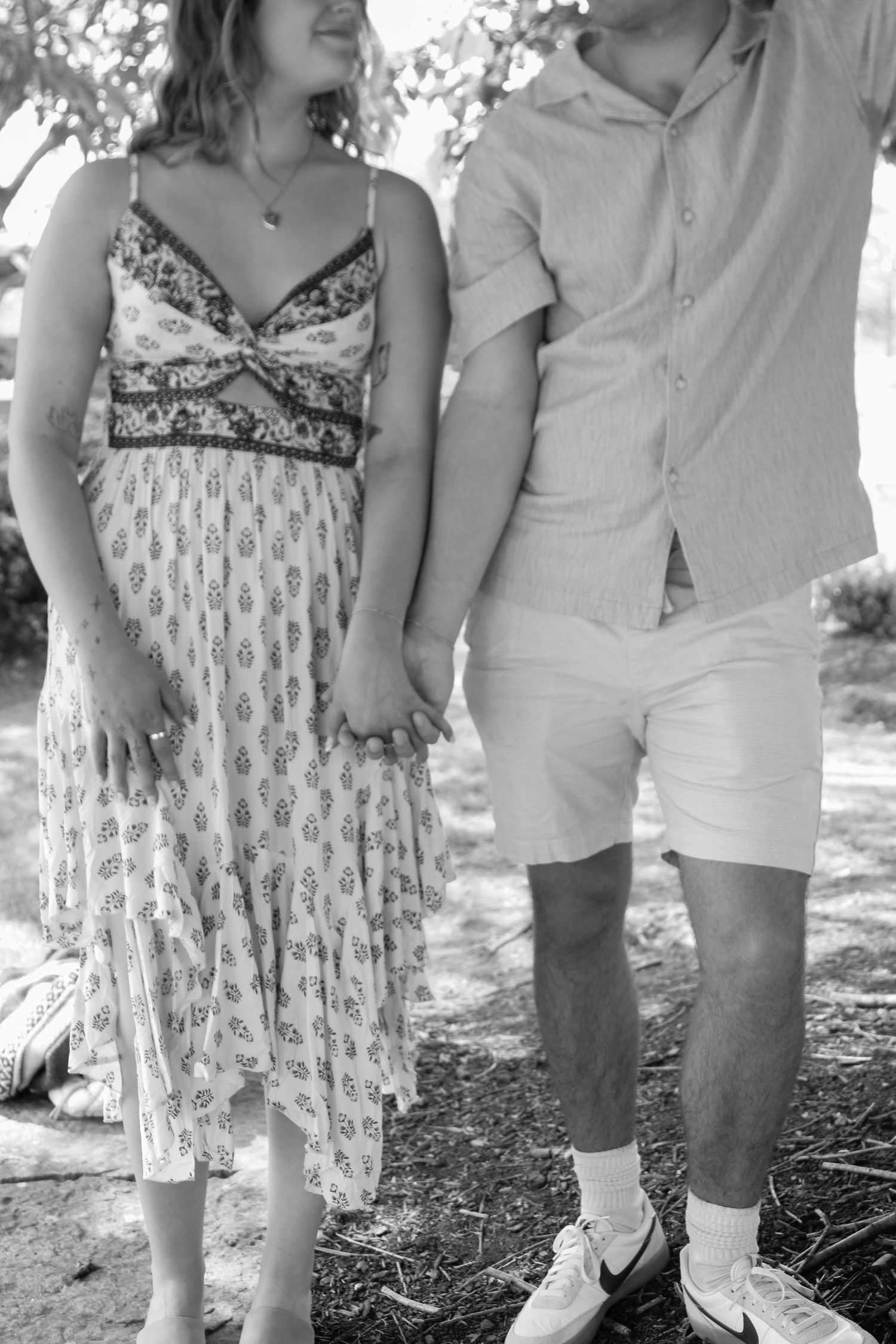A couple in summer attire stands together in dappled sunlight.