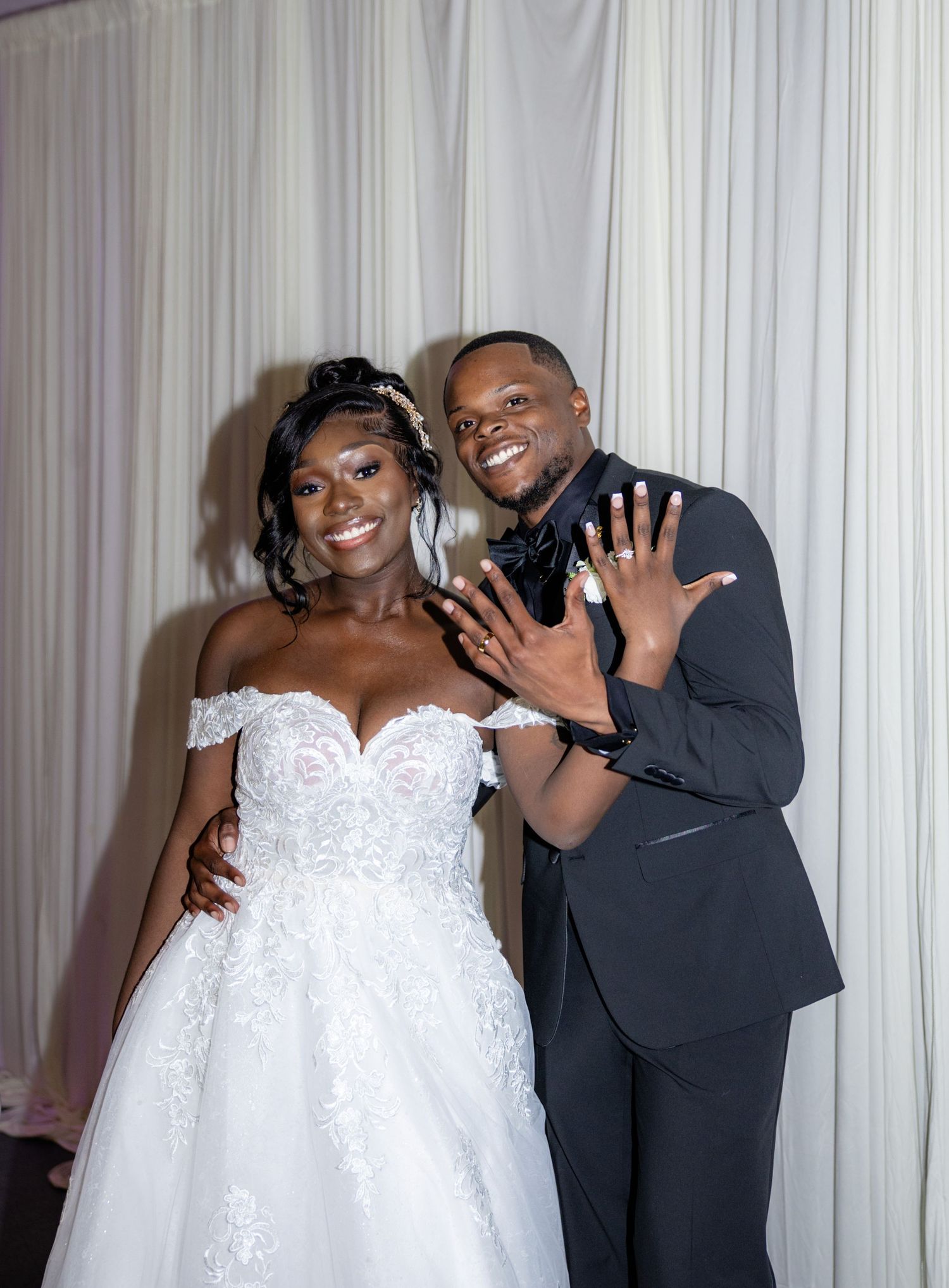 Bride and groom showing wedding rings, smiling in formal attire against white draped backdrop.