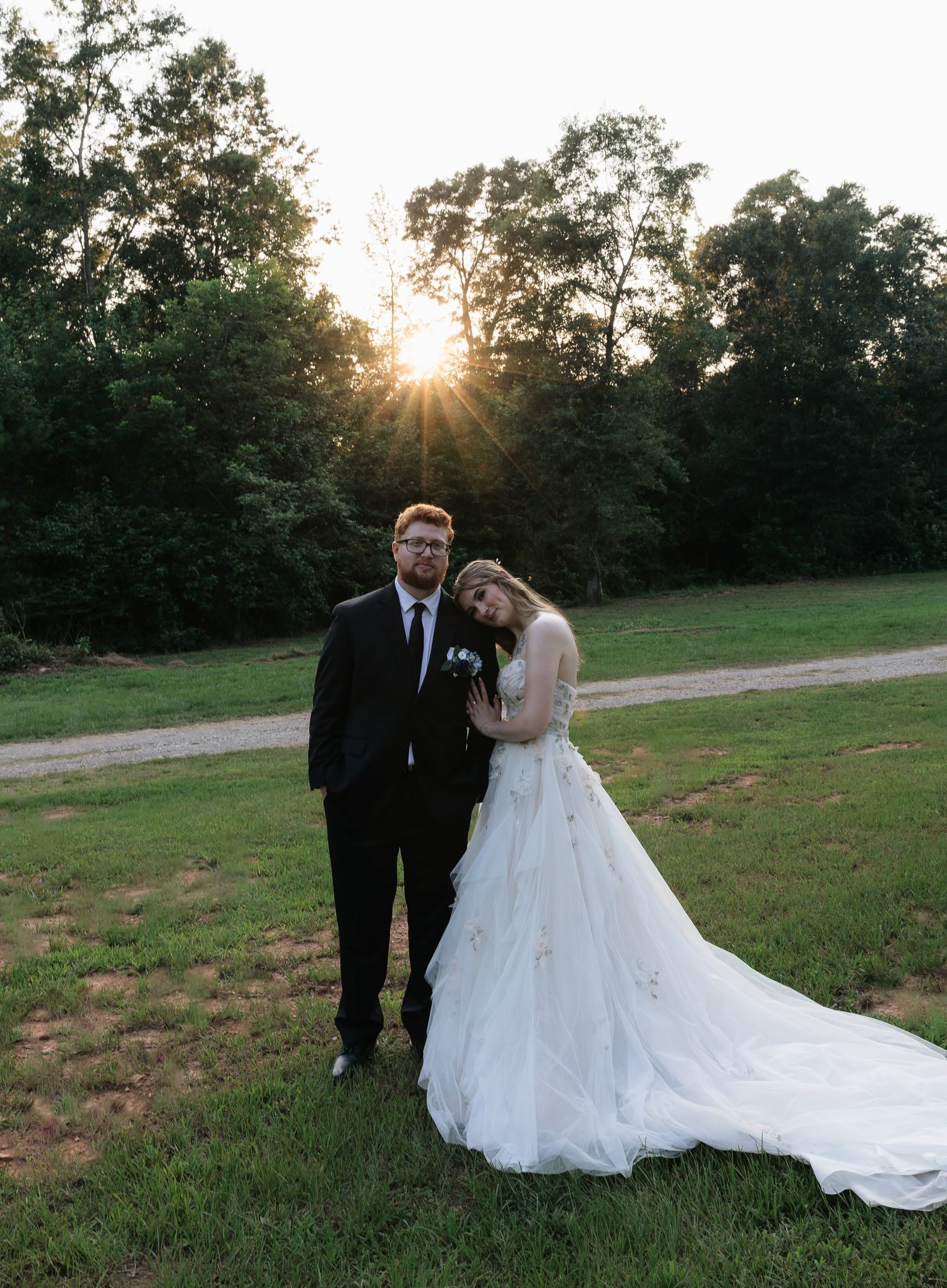 Bride and groom pose together at sunset in a field, bride in a flowing white gown with a long train.