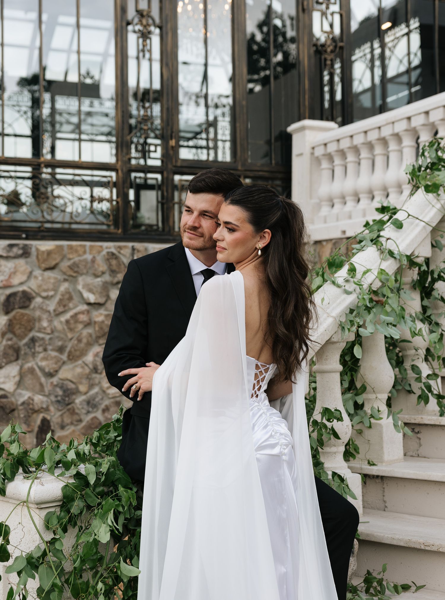 Bride in elegant white cape gown embraces groom in black suit on ivy-covered staircase at wedding venue.
