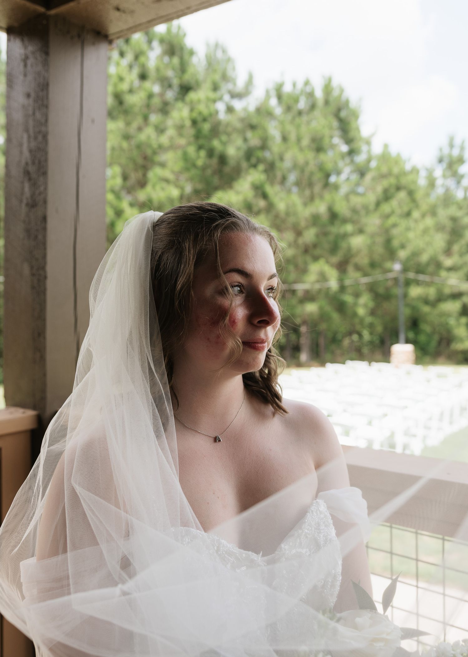 Bride in white wedding dress and veil gazing outdoors on wooden porch with lush green trees in background.