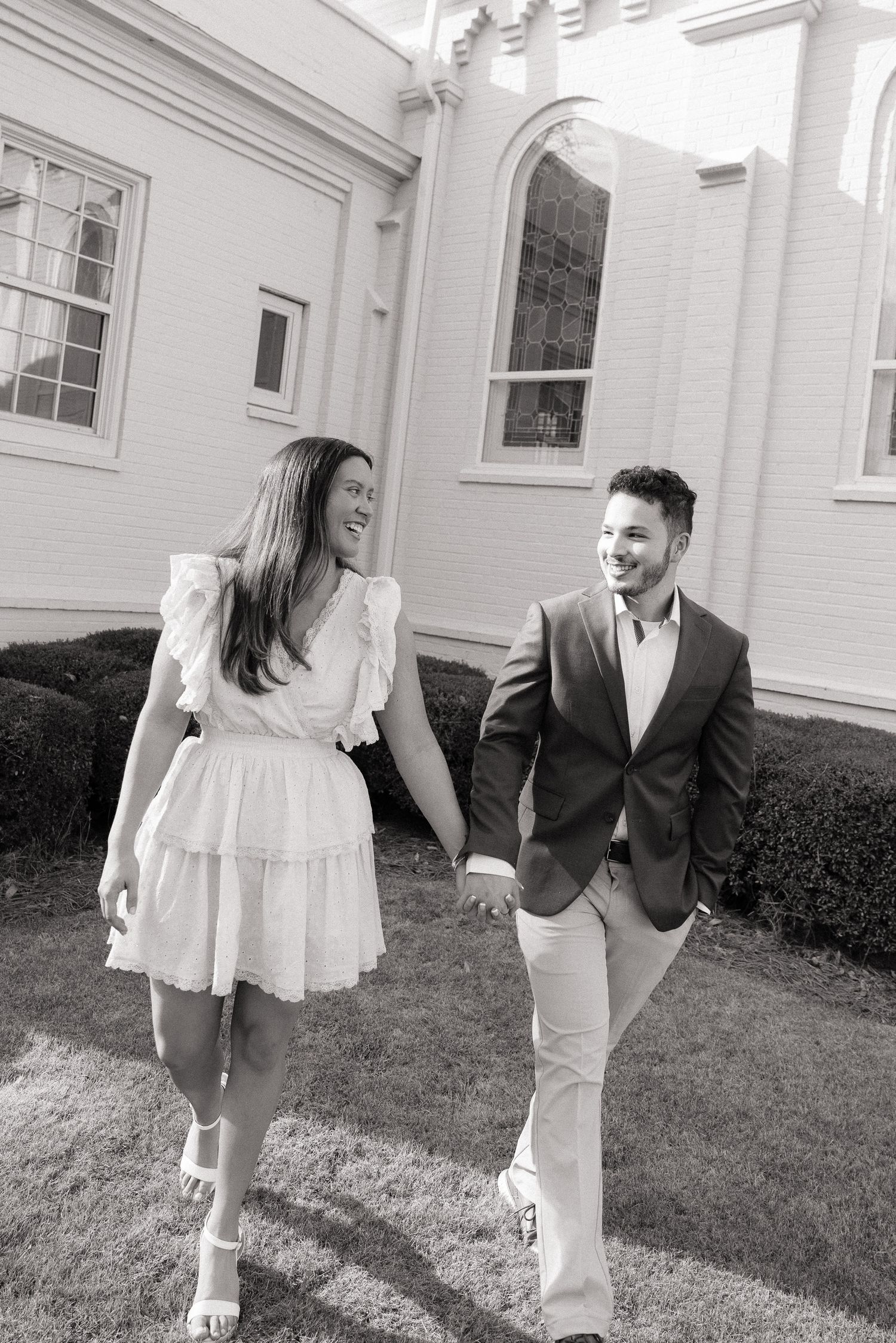 Black and white photo of a smiling couple holding hands walking outside a church building.