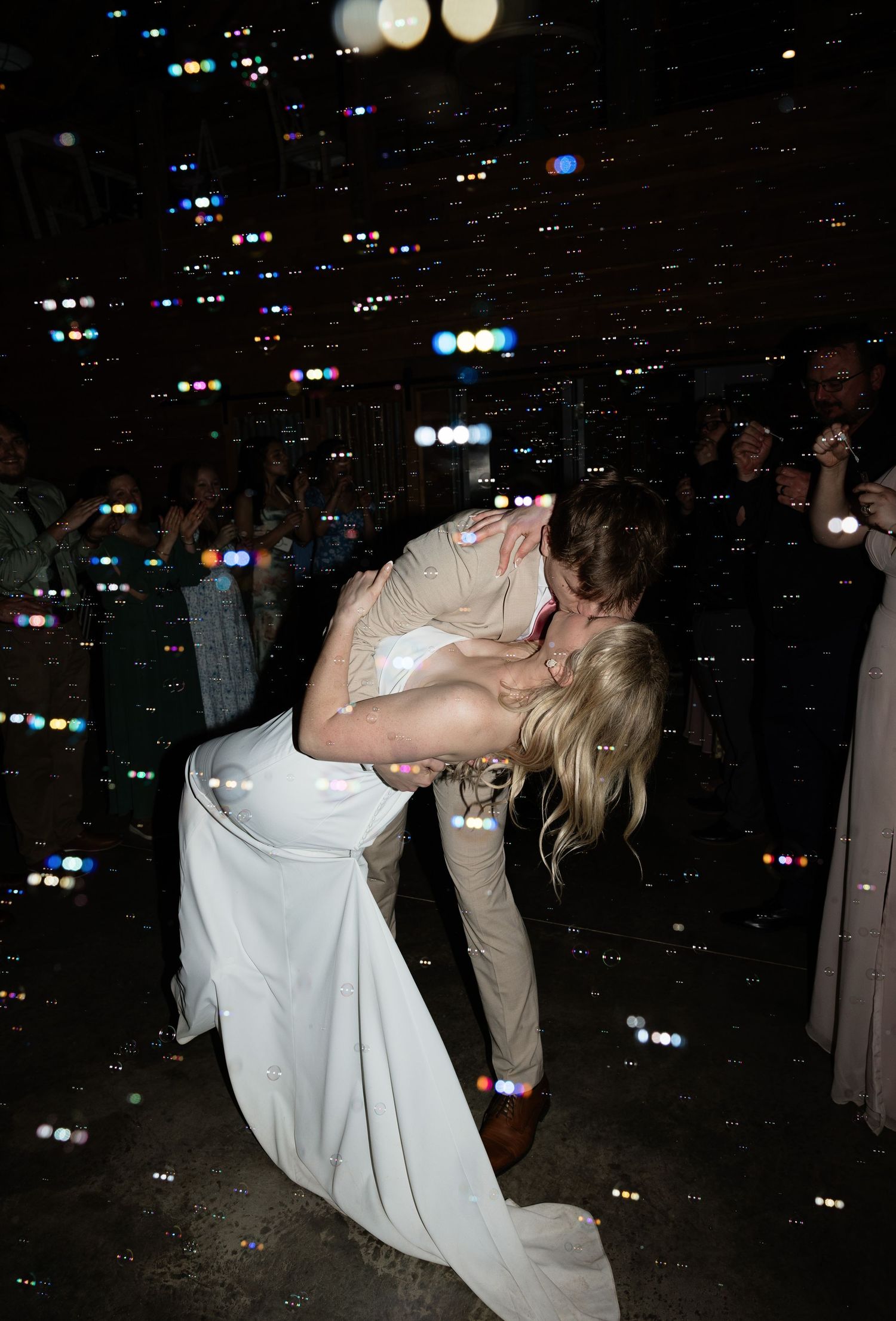 Bride and groom share a romantic dip kiss on the dance floor surrounded by bubbles and guests at their wedding reception.