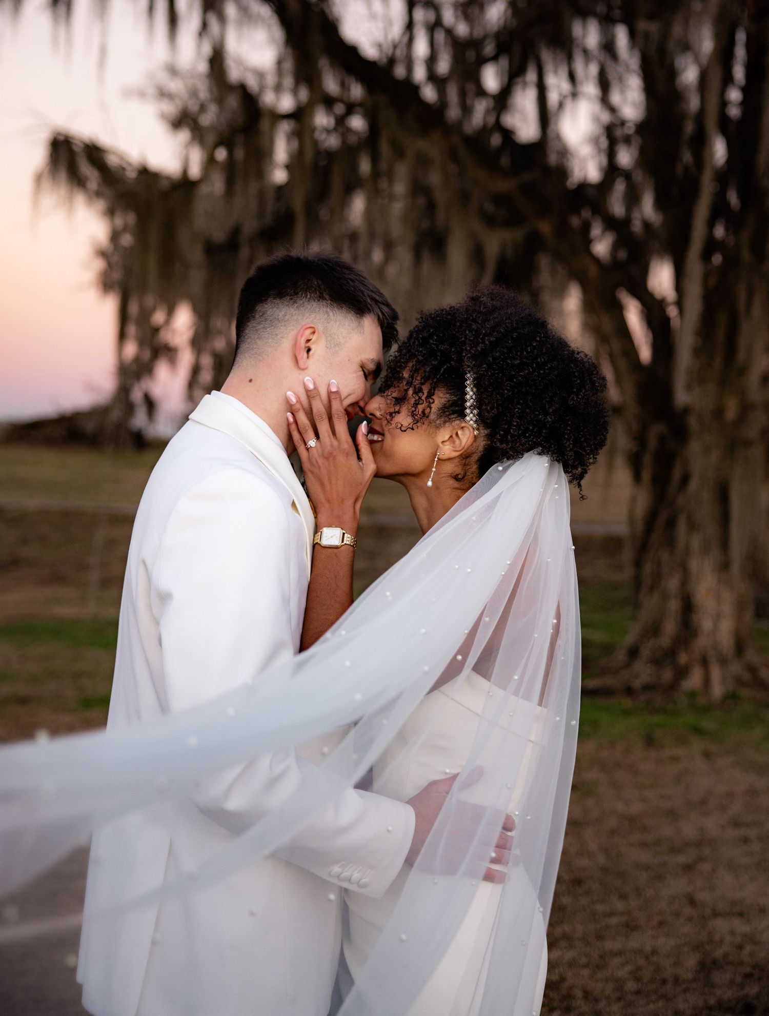 Newlywed couple shares intimate moment under Spanish moss trees at sunset with flowing white veil and attire.