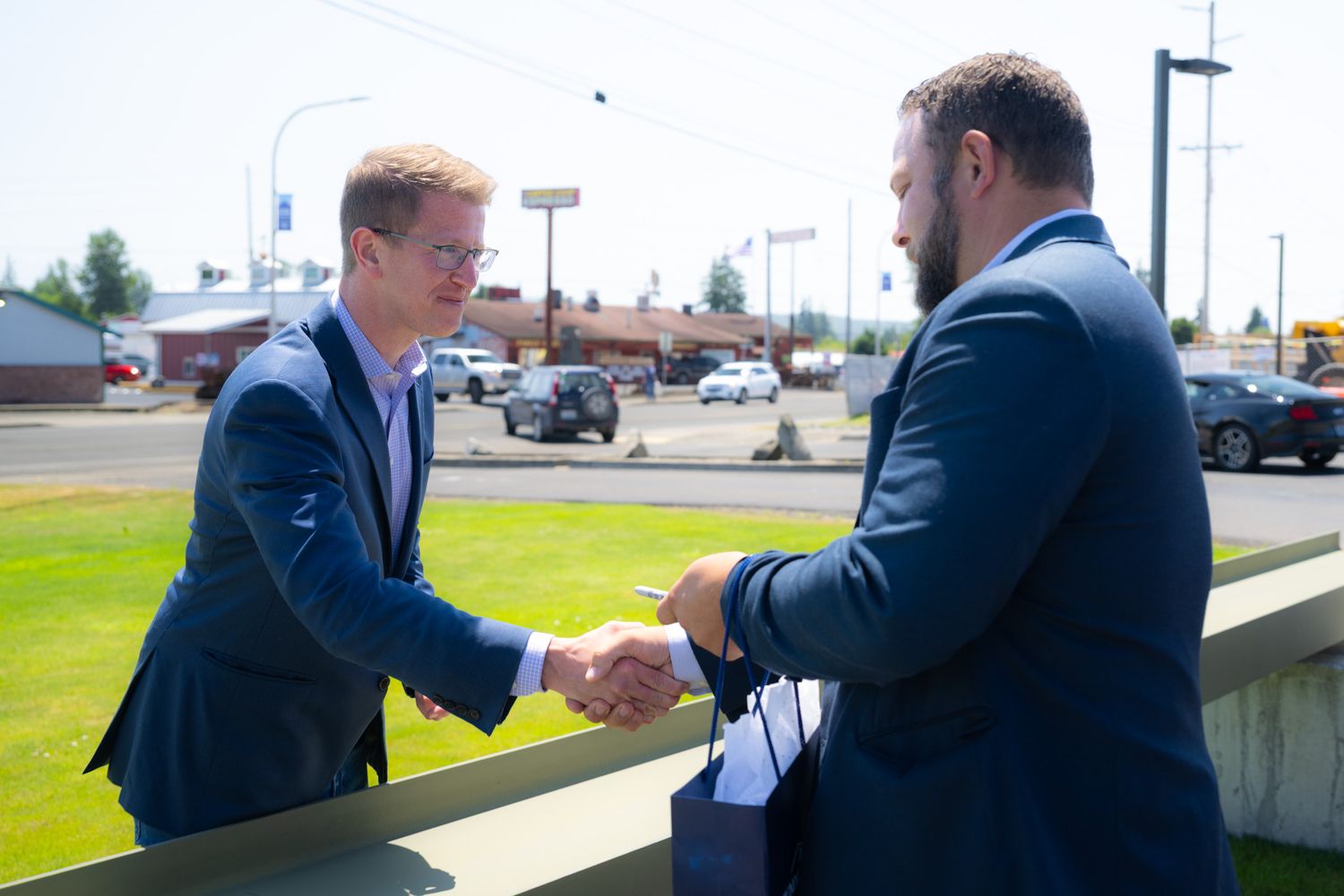 Two men in dark suits shake hands outdoors on a sunny day beside a low wall, with a parking lot and trees in the background.
