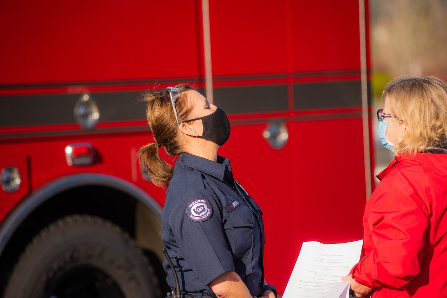 A masked firefighter in a navy uniform looks upward beside a red fire truck while a woman in a red jacket holds papers nearby.