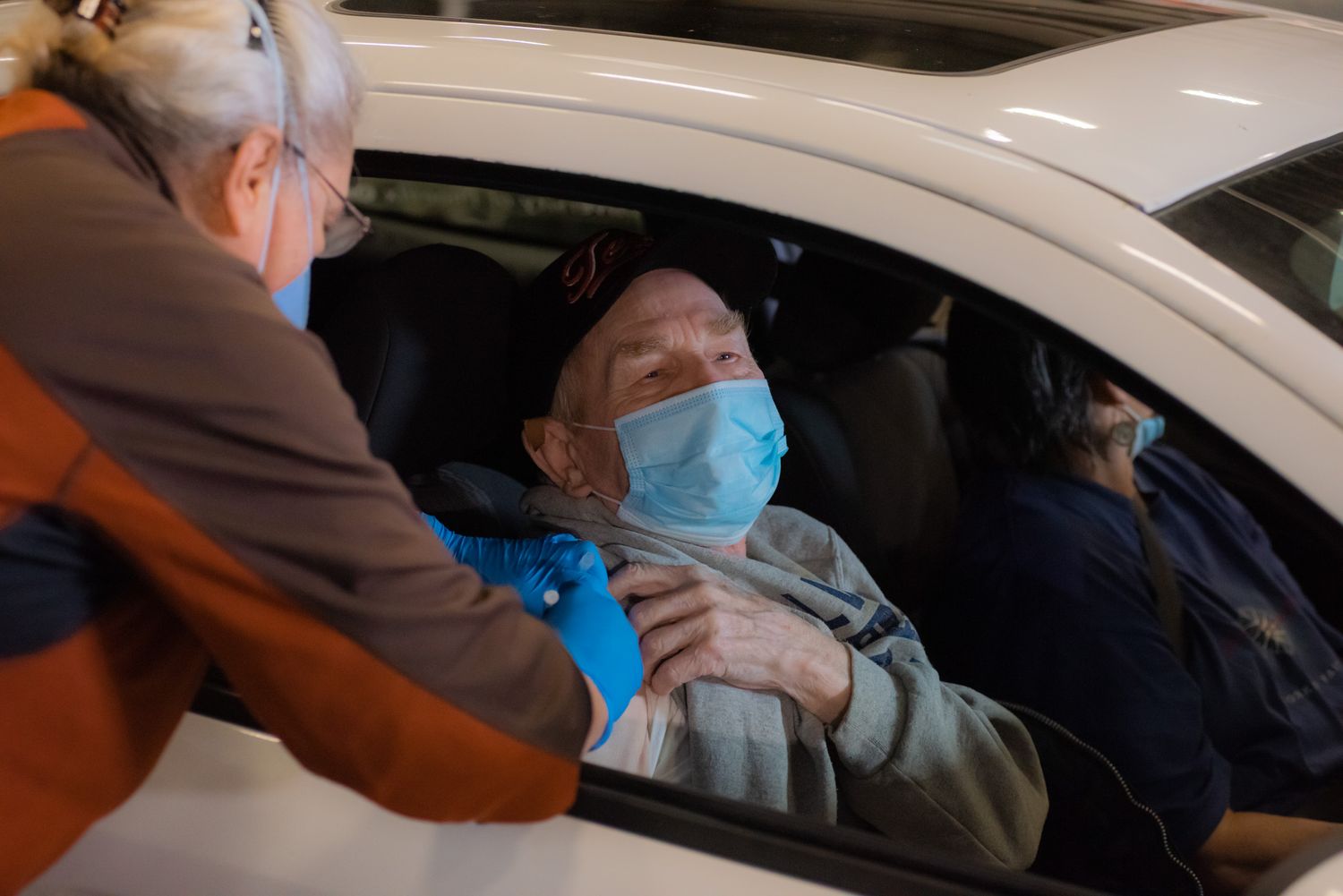Elderly man in a blue face mask receives a vaccine shot in his arm from a healthcare worker while seated in a car at a drive-through clinic.