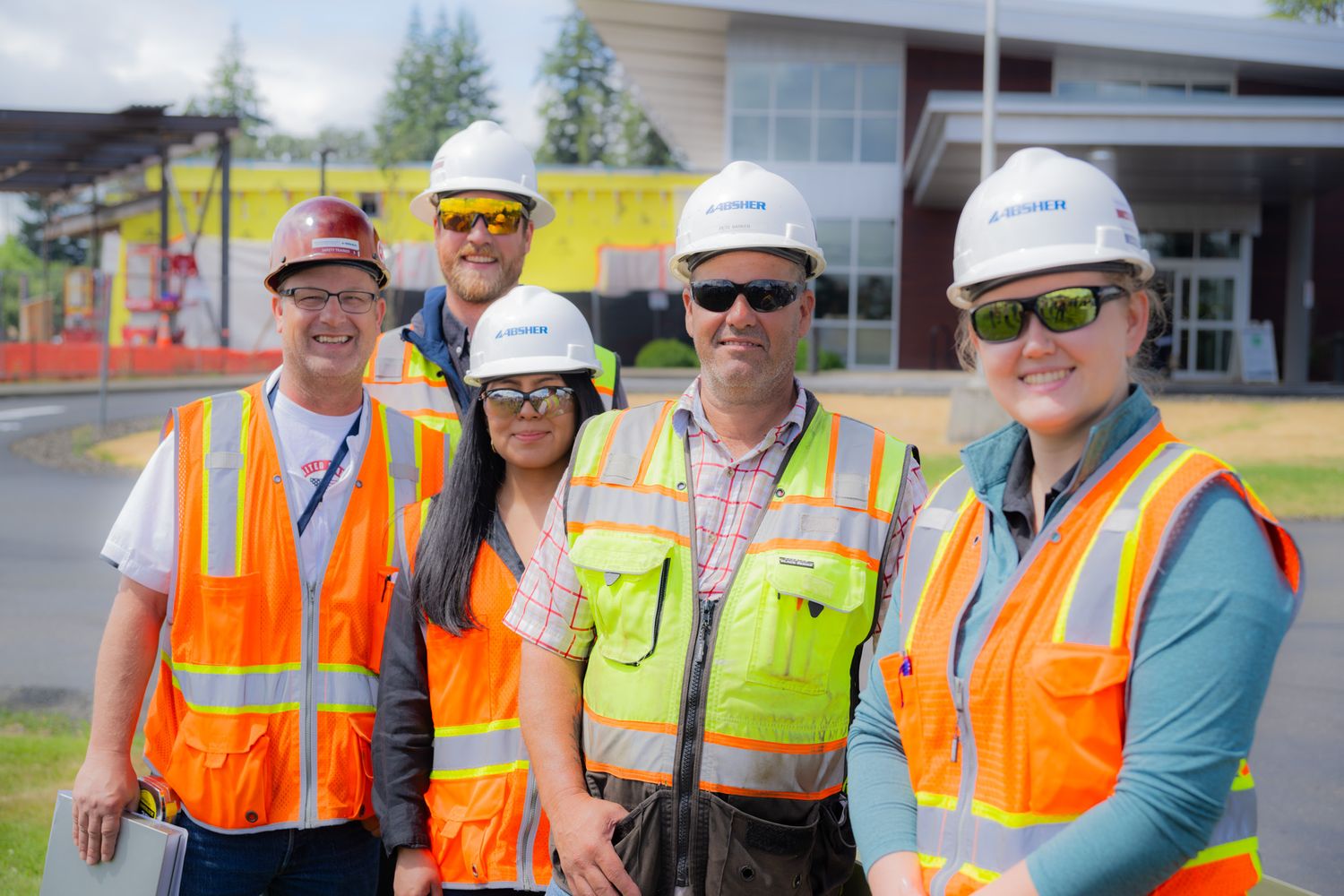 Five smiling construction workers in hard hats and high-visibility safety vests pose together outdoors in front of a modern building.