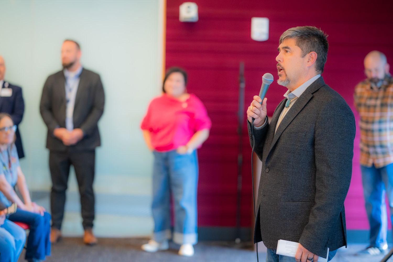 A man in a dark blazer speaks into a microphone at a community event while attendees stand and sit in the background.