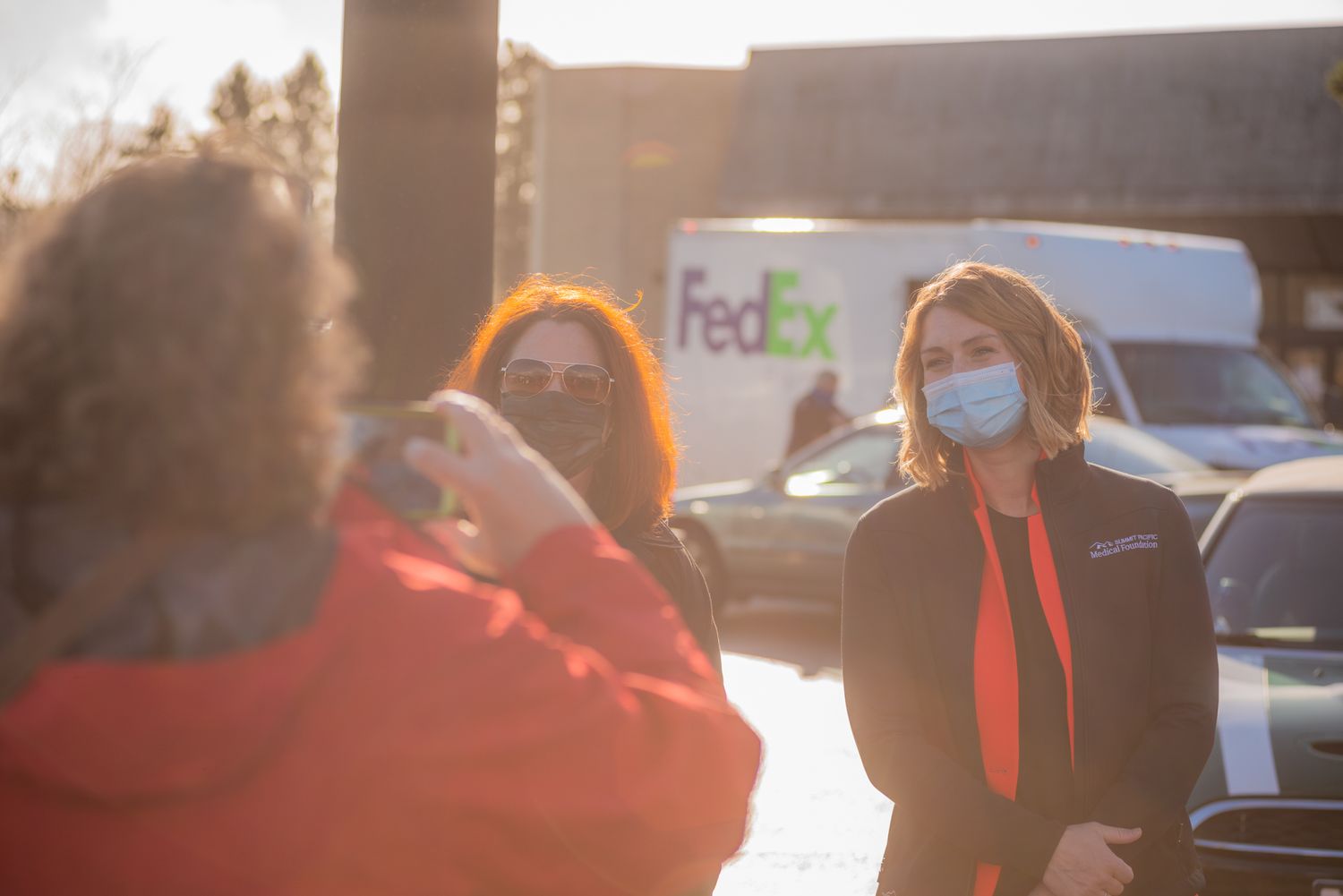Woman in a blue face mask and orange scarf stands in a parking lot while others take photos, with a FedEx truck behind.